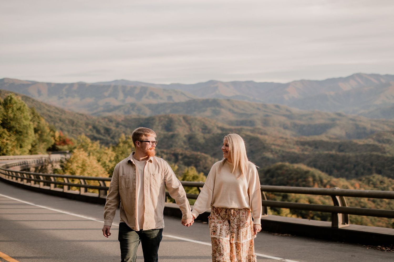 A couple walks hand in hand on a scenic mountain overlook during golden hour with rolling hills in the background.