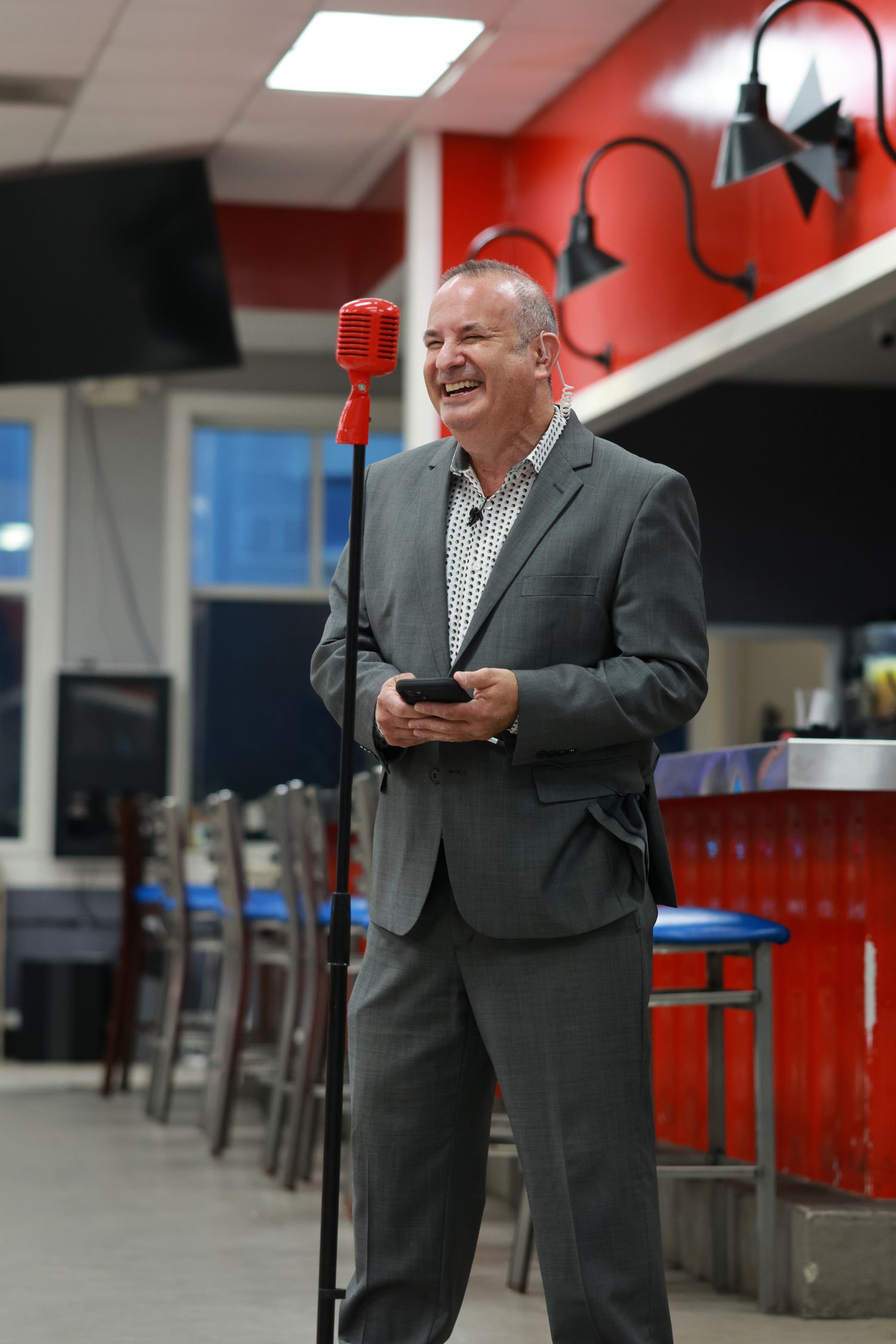 A speaker in a grey suit holds a mobile device while standing at a microphone in a casual venue.