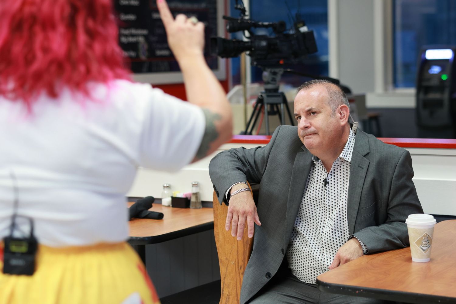 A person with red hair interviews someone in a grey suit at a desk with recording equipment.