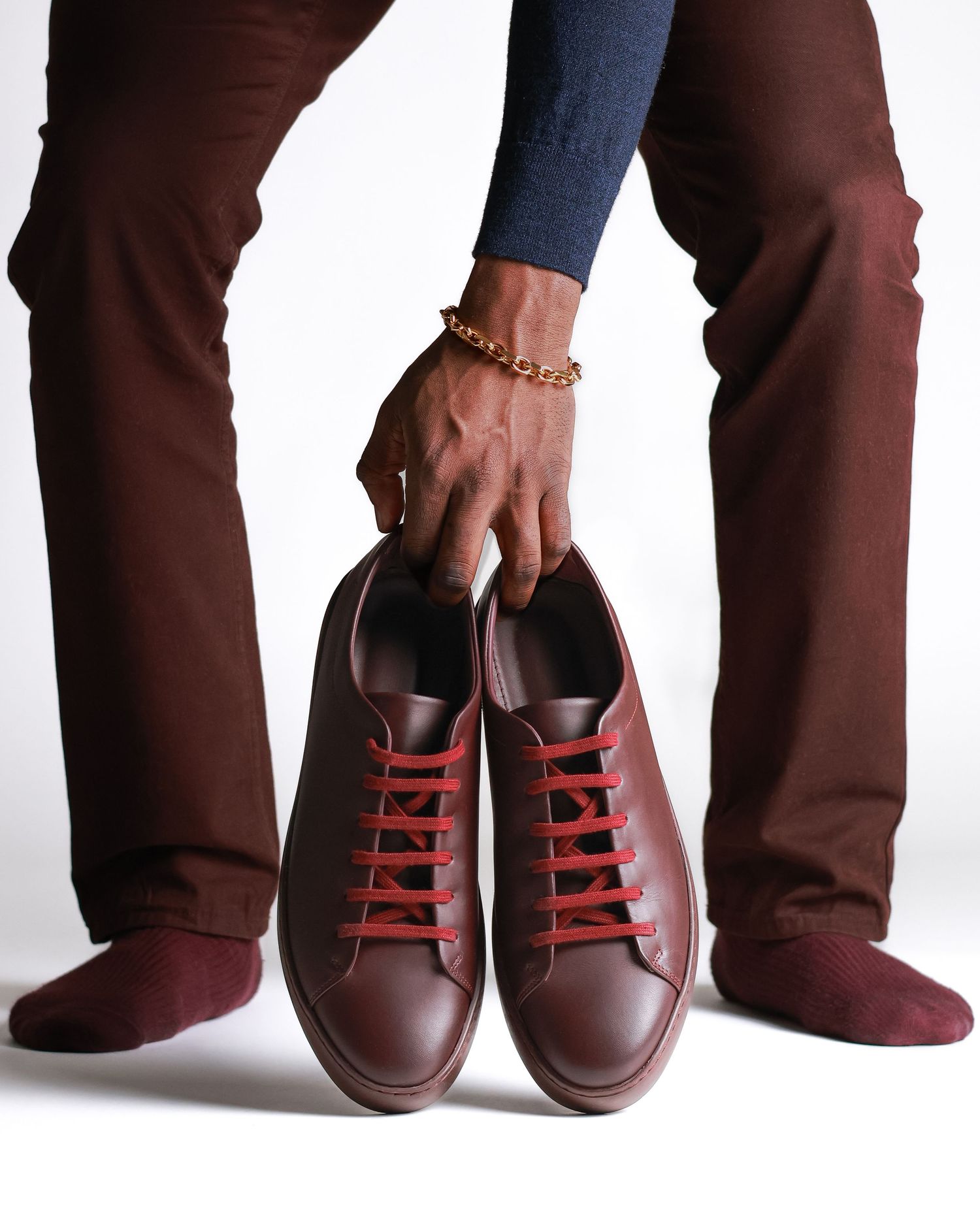 Person holding brown leather sneakers with red laces while wearing burgundy pants.