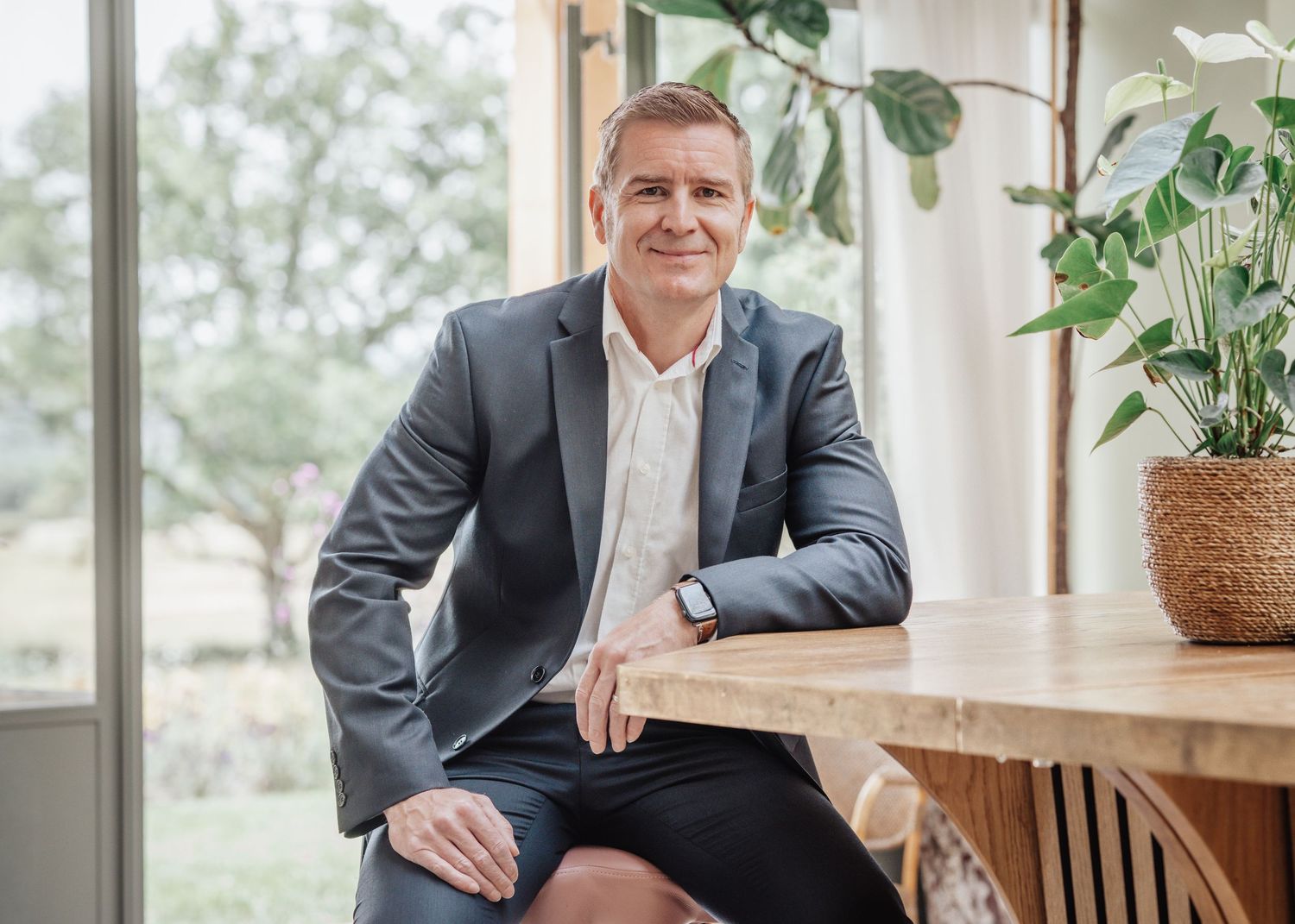 Professional man in gray blazer smiling, seated at wooden table in modern office with green plants.