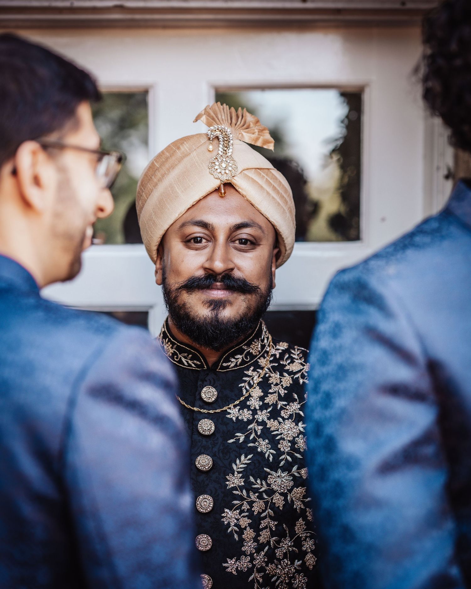 Indian groom in traditional black sherwani with gold embroidery and cream turban smiling at wedding ceremony.