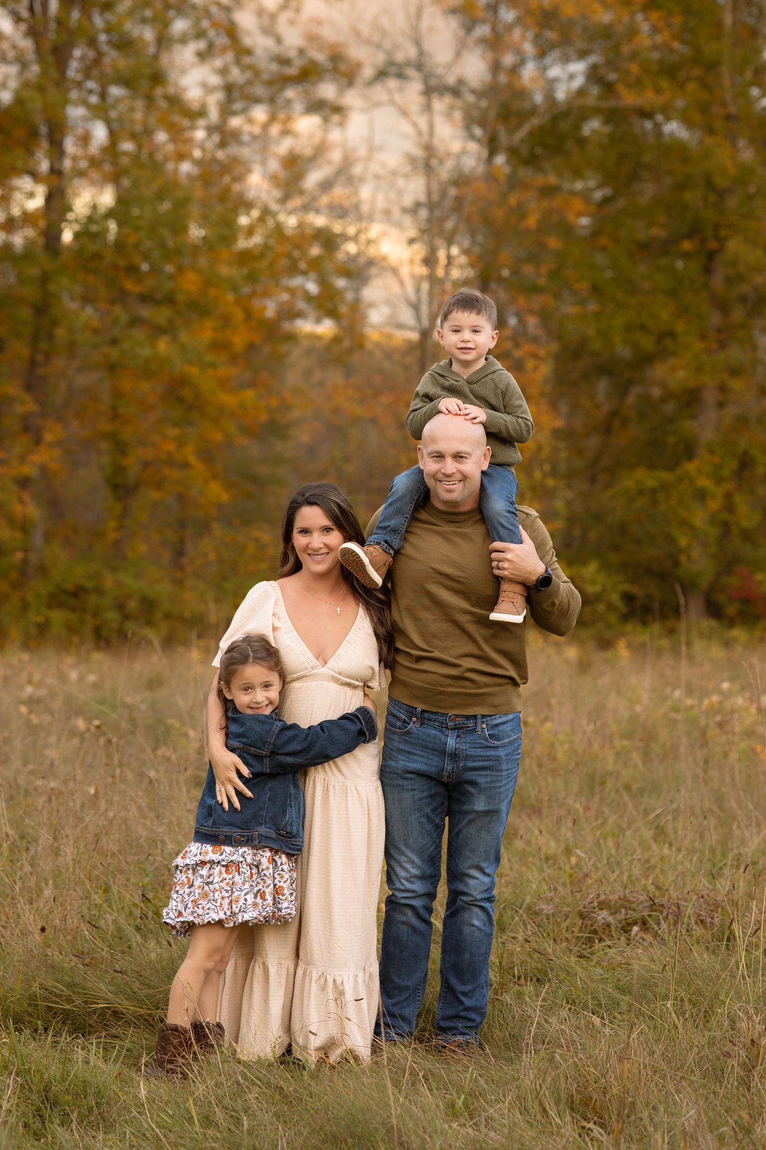 Family of four posing in autumn field, mom in cream dress, dad with son on shoulders, daughter hugging mom.