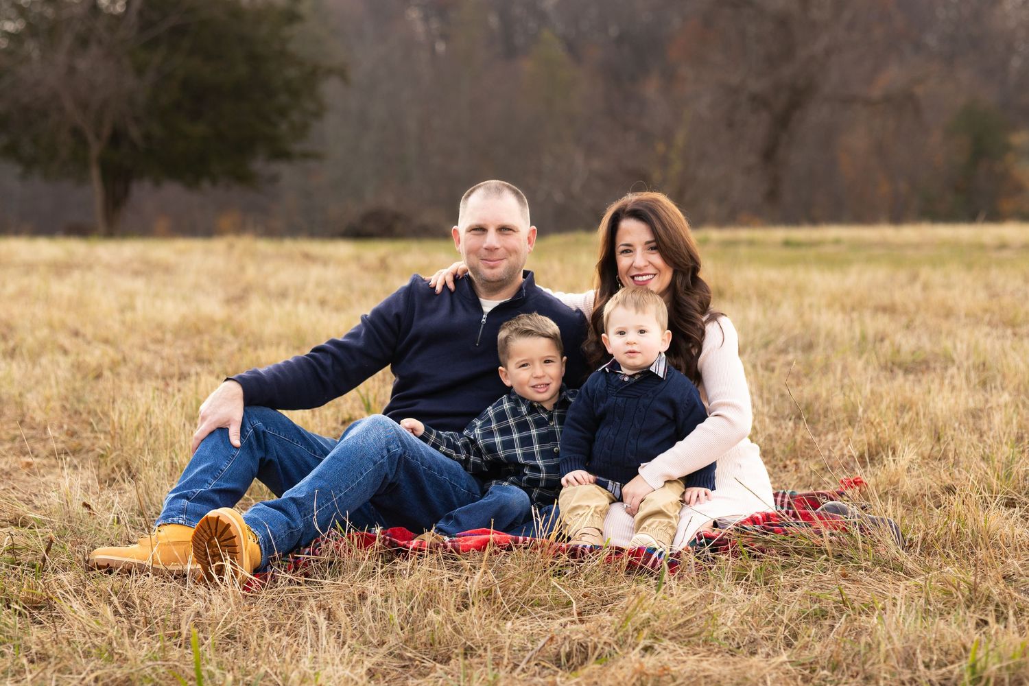 Family of four smiling together on a red blanket in an open field during a fall outdoor photo session.