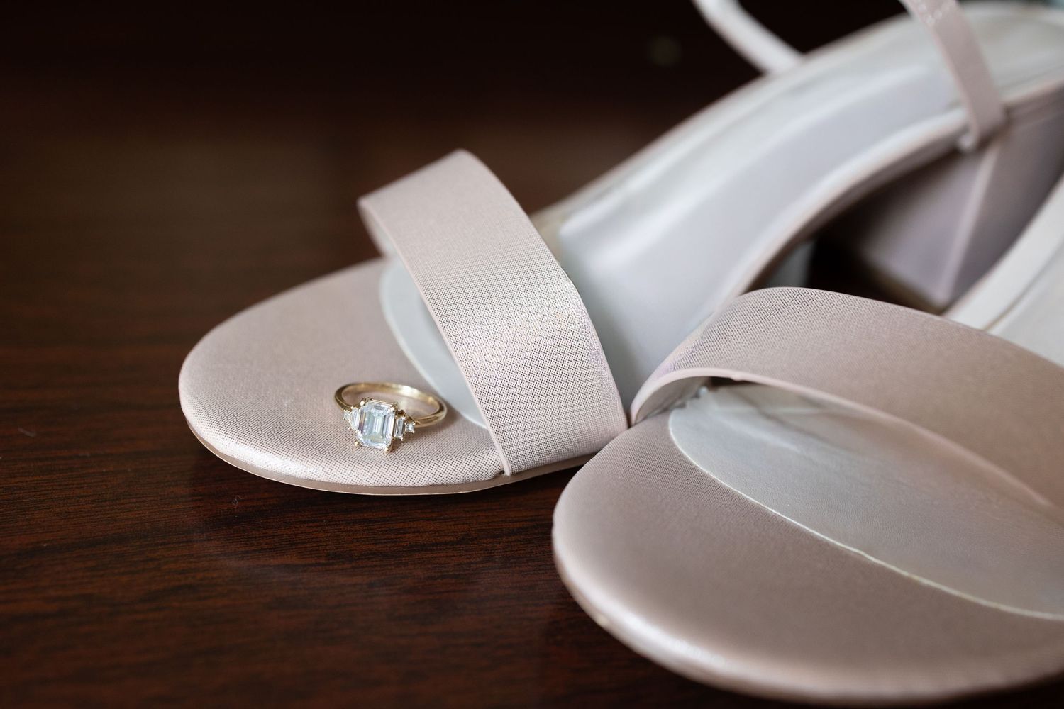 White bridal shoes with delicate straps arranged on dark wooden surface. Wedding. Rings