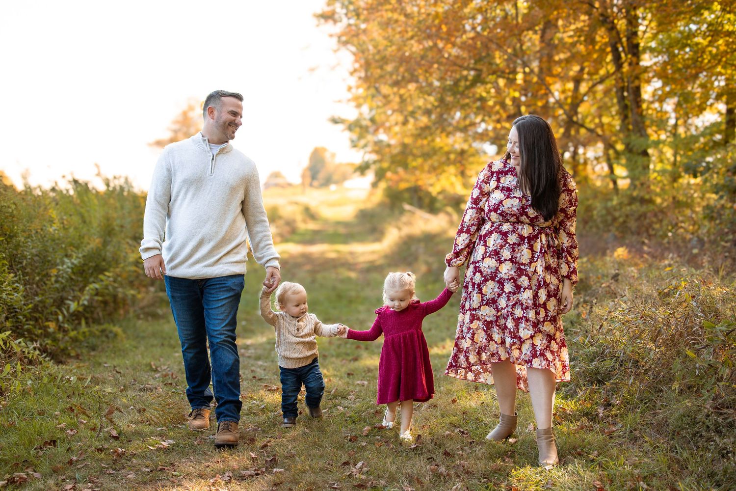 Family of four walking hand-in-hand on a sunlit autumn trail surrounded by golden foliage.