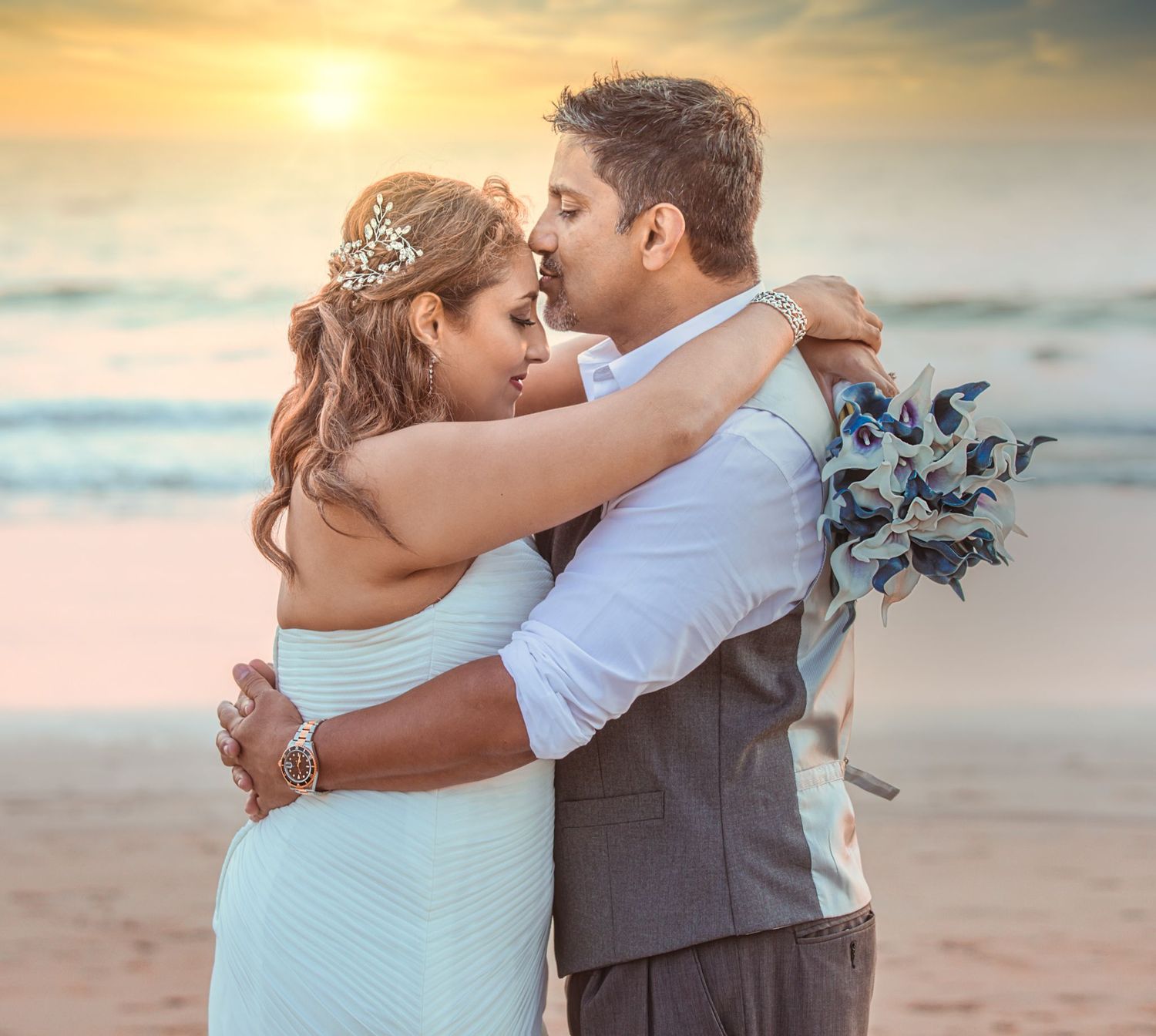 A romantic couple embraces on a sandy beach at sunset with soft golden light illuminating the scene.