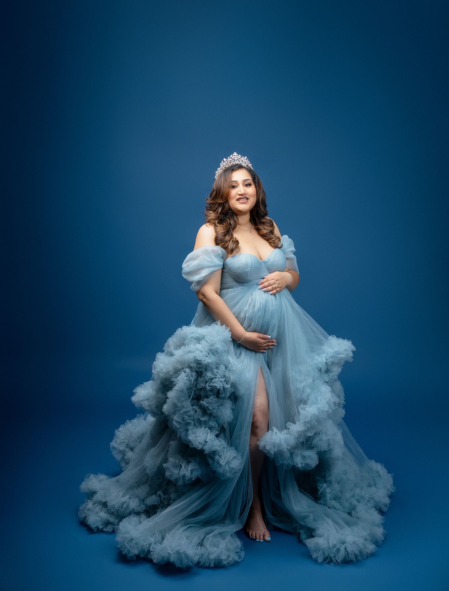 A maternity photoshoot featuring a flowing powder blue tulle gown against a dark blue studio backdrop.