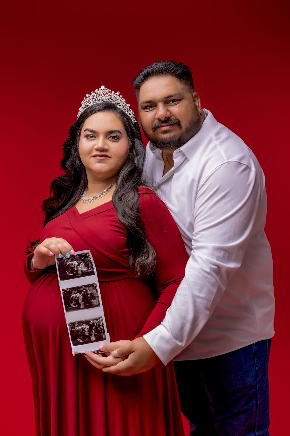 A couple in red and white attire poses with ultrasound photos against a deep red backdrop during a pregnancy photoshoot.