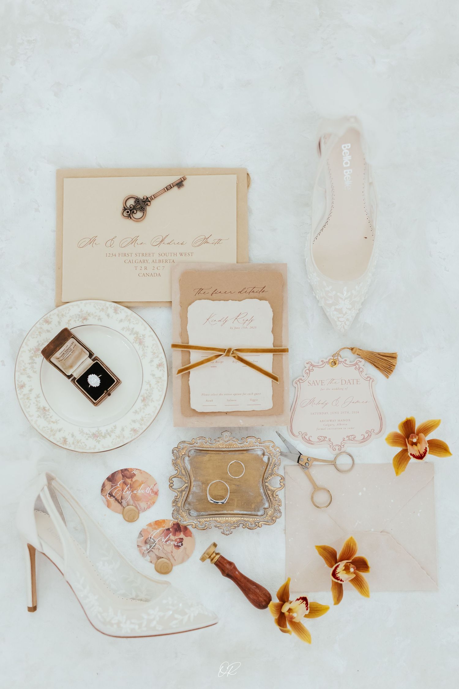 Elegant wedding accessories flatlay with white heels, ring box, invitations, and orange orchids on light background.