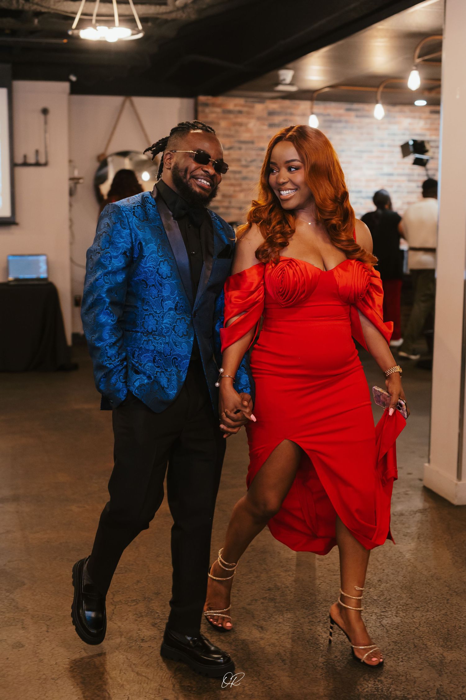 A couple dressed in formal attire, with a royal blue blazer and red off-shoulder dress, dancing together in an indoor venue.