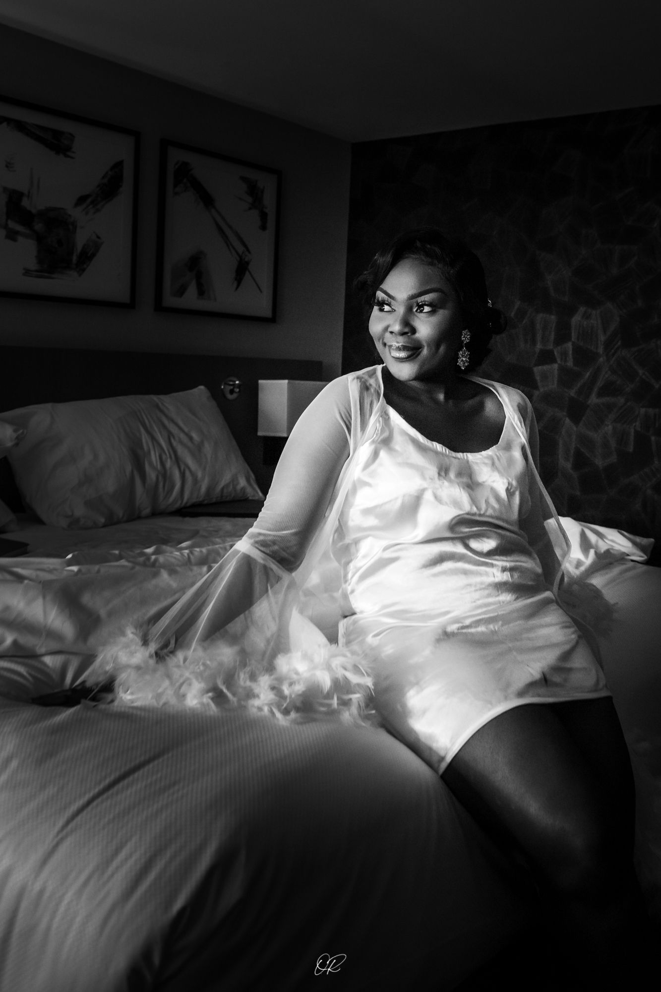 Black and white boudoir photograph showing a person in white sleepwear relaxing on a bed with wall art visible in background.