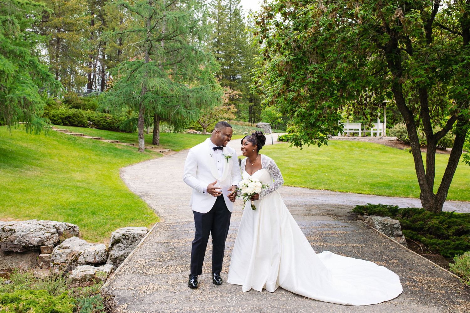 A couple in wedding attire walks together on a winding path through a lush green garden surrounded by trees.