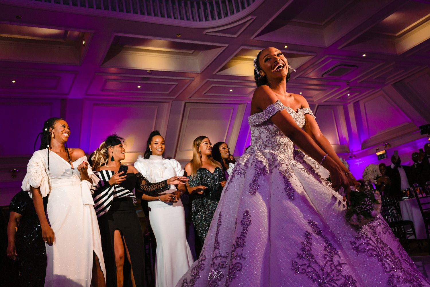 Wedding reception dance party with guests and a bride in a white ballgown under purple lighting in an elegant venue.