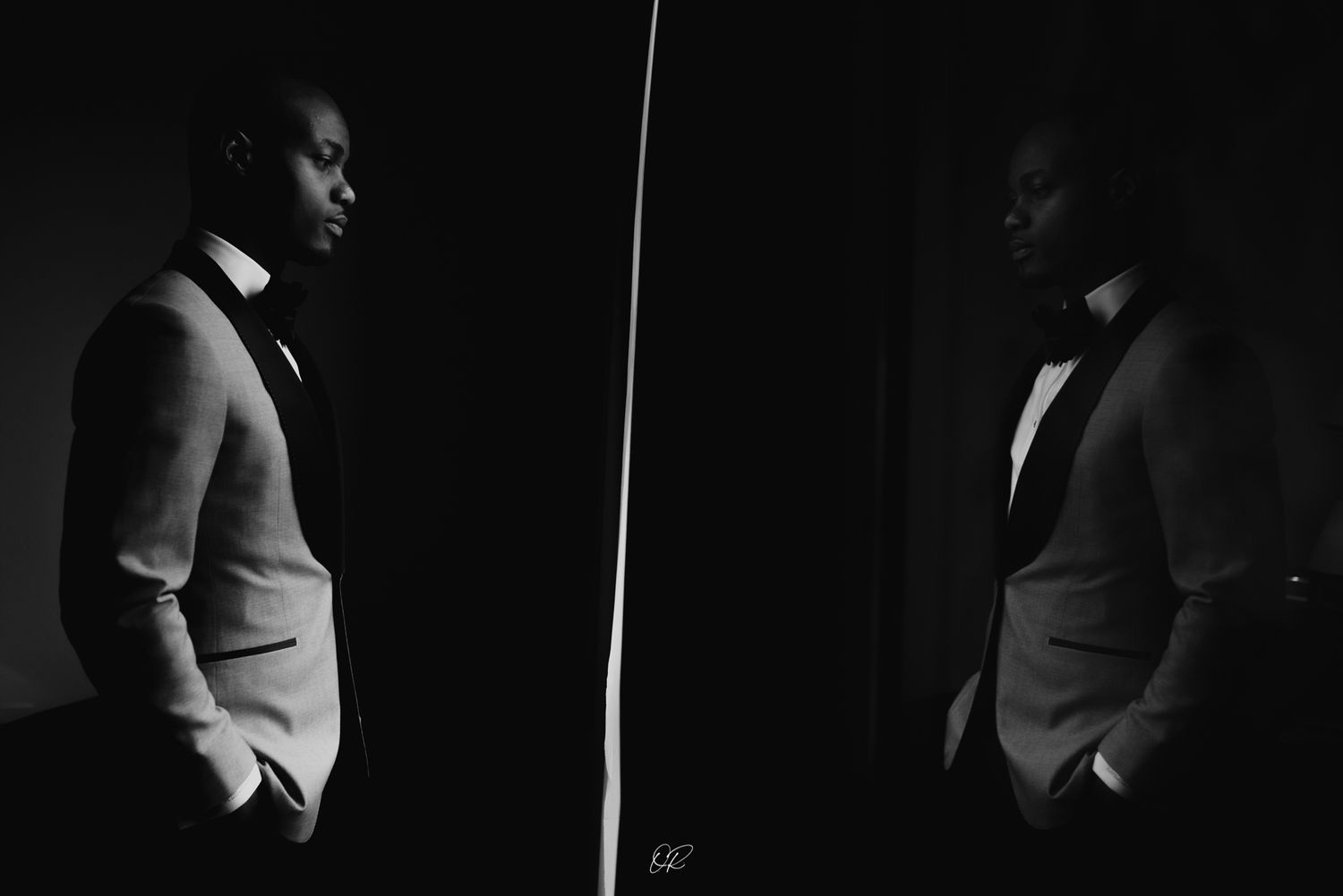 A dramatic black and white portrait of a figure in formal attire standing beside a mirror reflection in low-key lighting.
