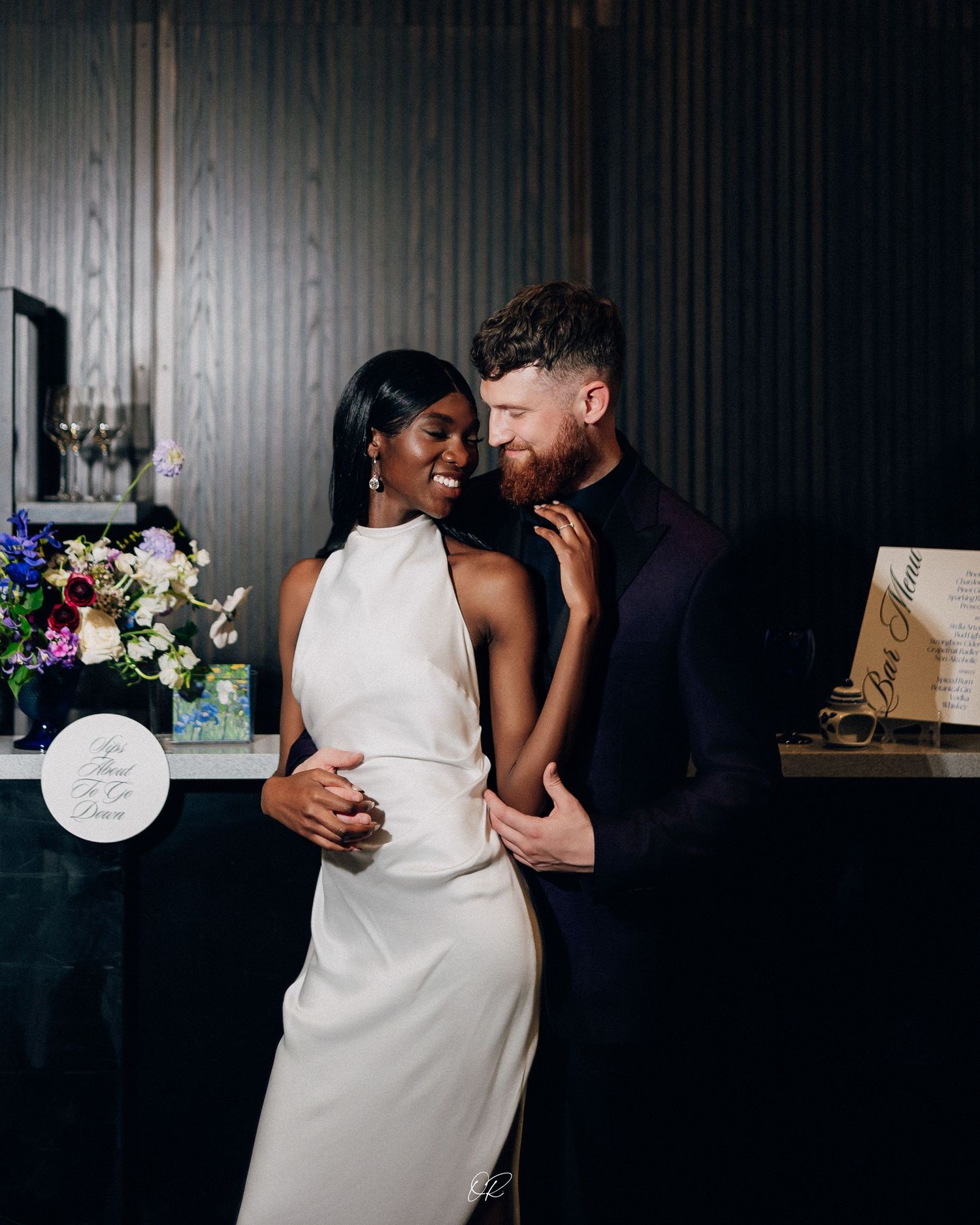 Intimate portrait of couple embracing in formal attire against dark background with floral decor.