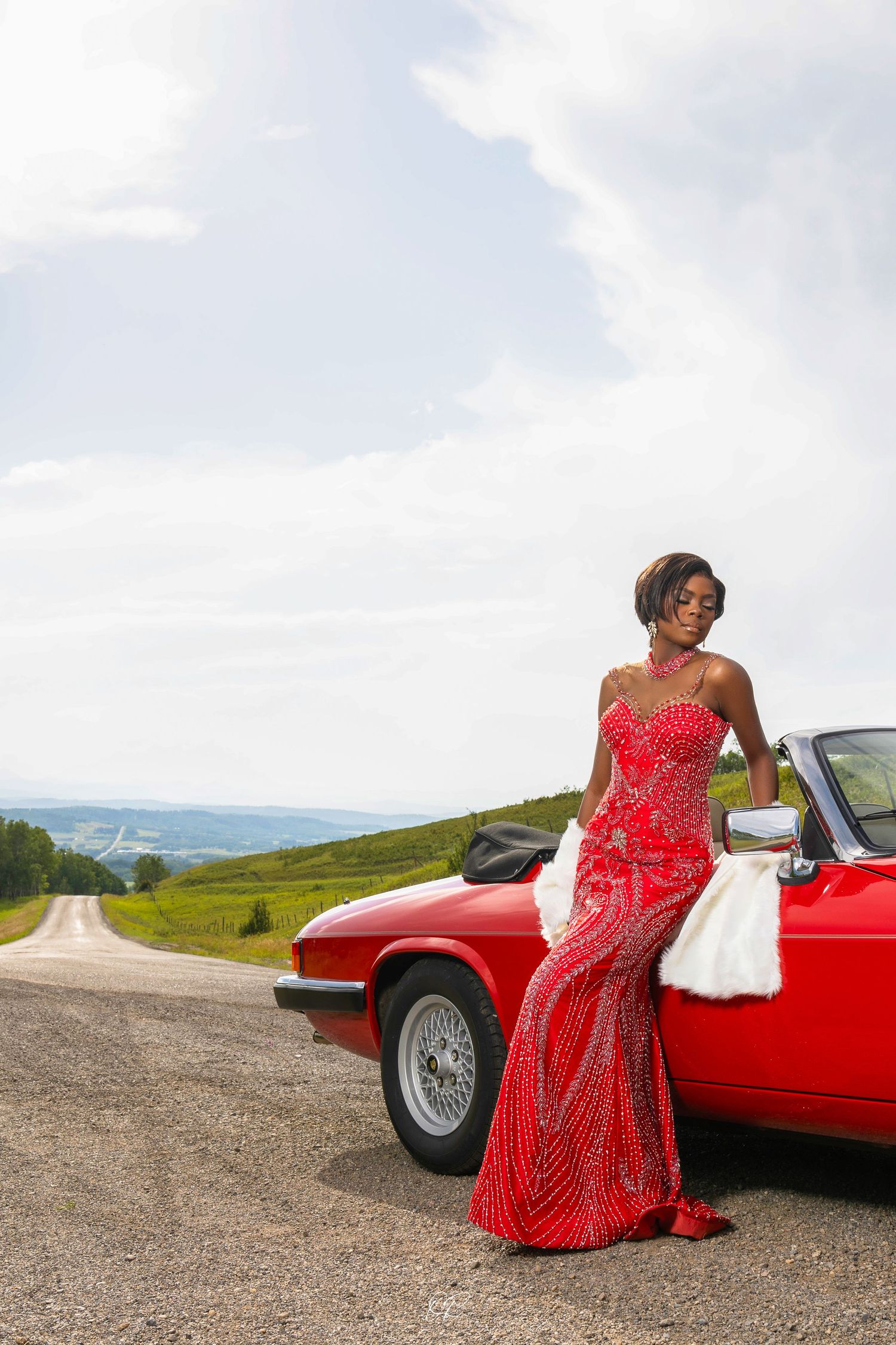 Red vintage car parked on scenic countryside road with mountains in background.