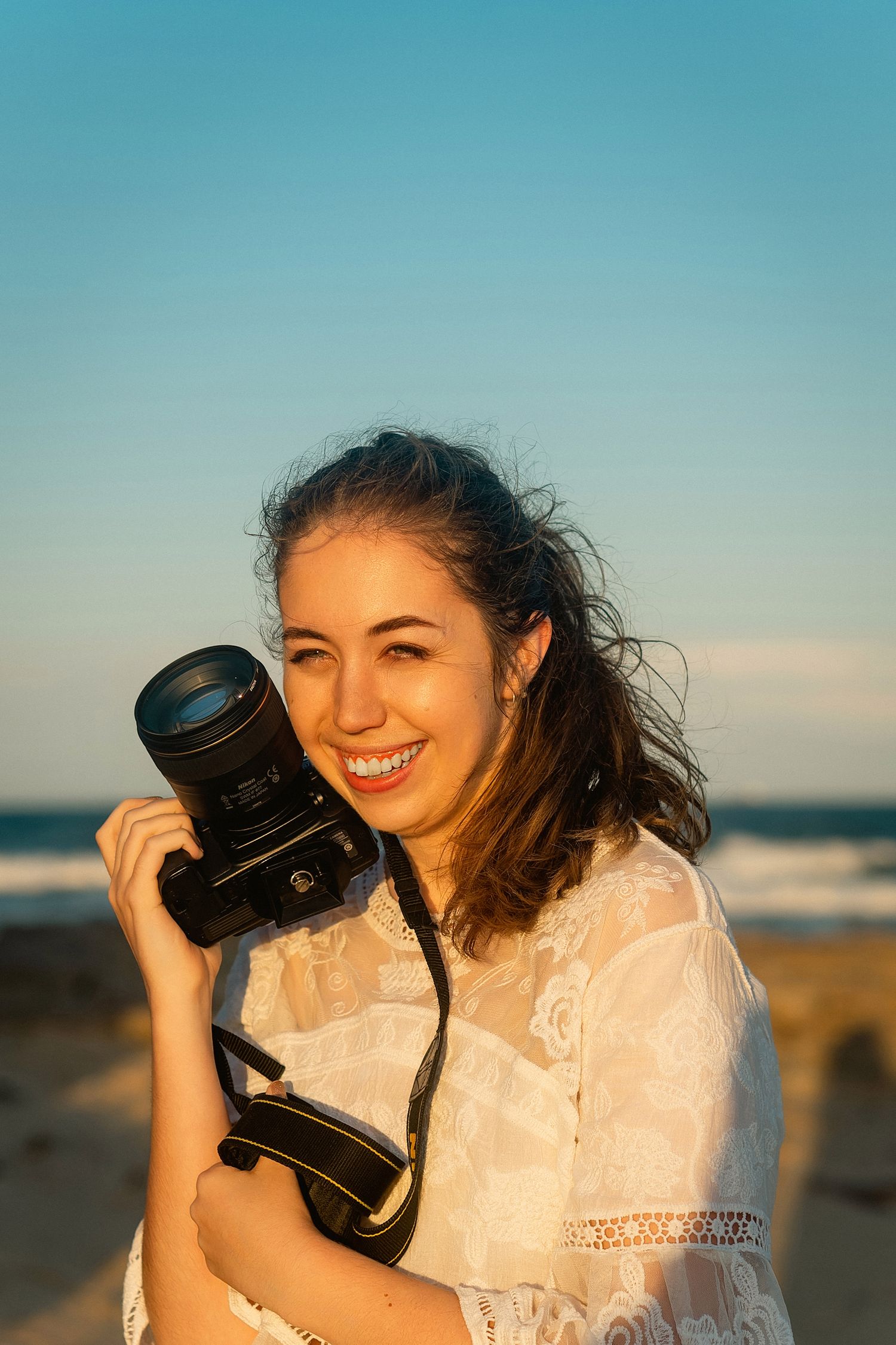 A photographer in a white lace dress holds a camera at sunset on the beach with golden light and ocean in background.
