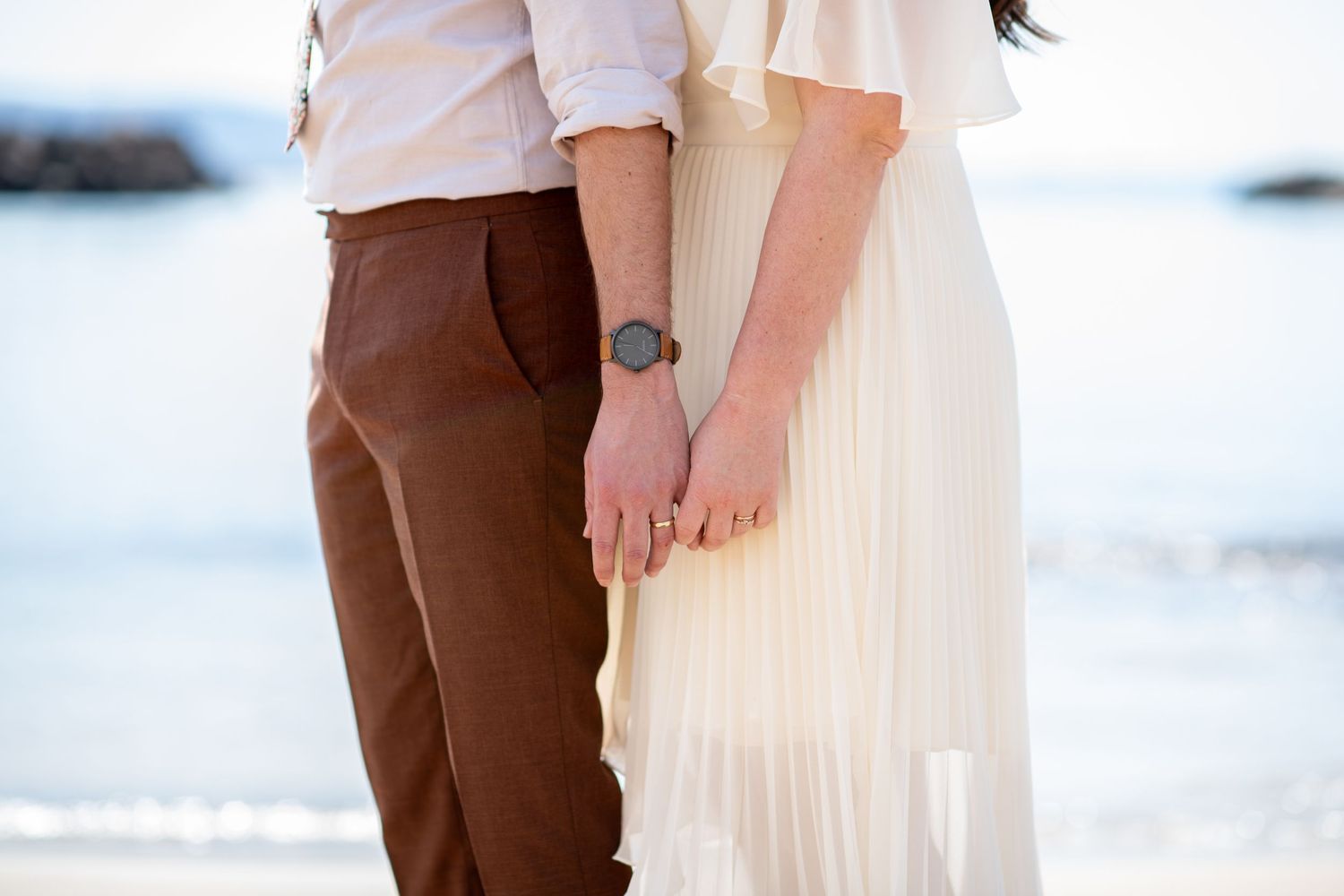 A couple holds hands on a beach at sunset wearing white and brown casual attire.