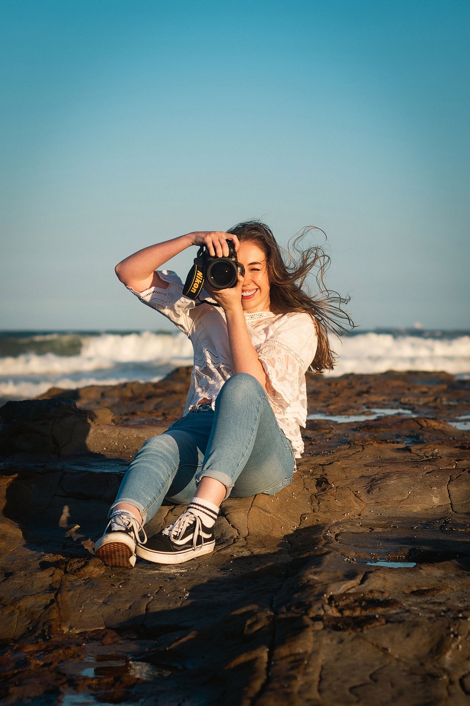 A person in casual attire lounges on rocky coastal terrain at sunset with ocean waves crashing in the background.