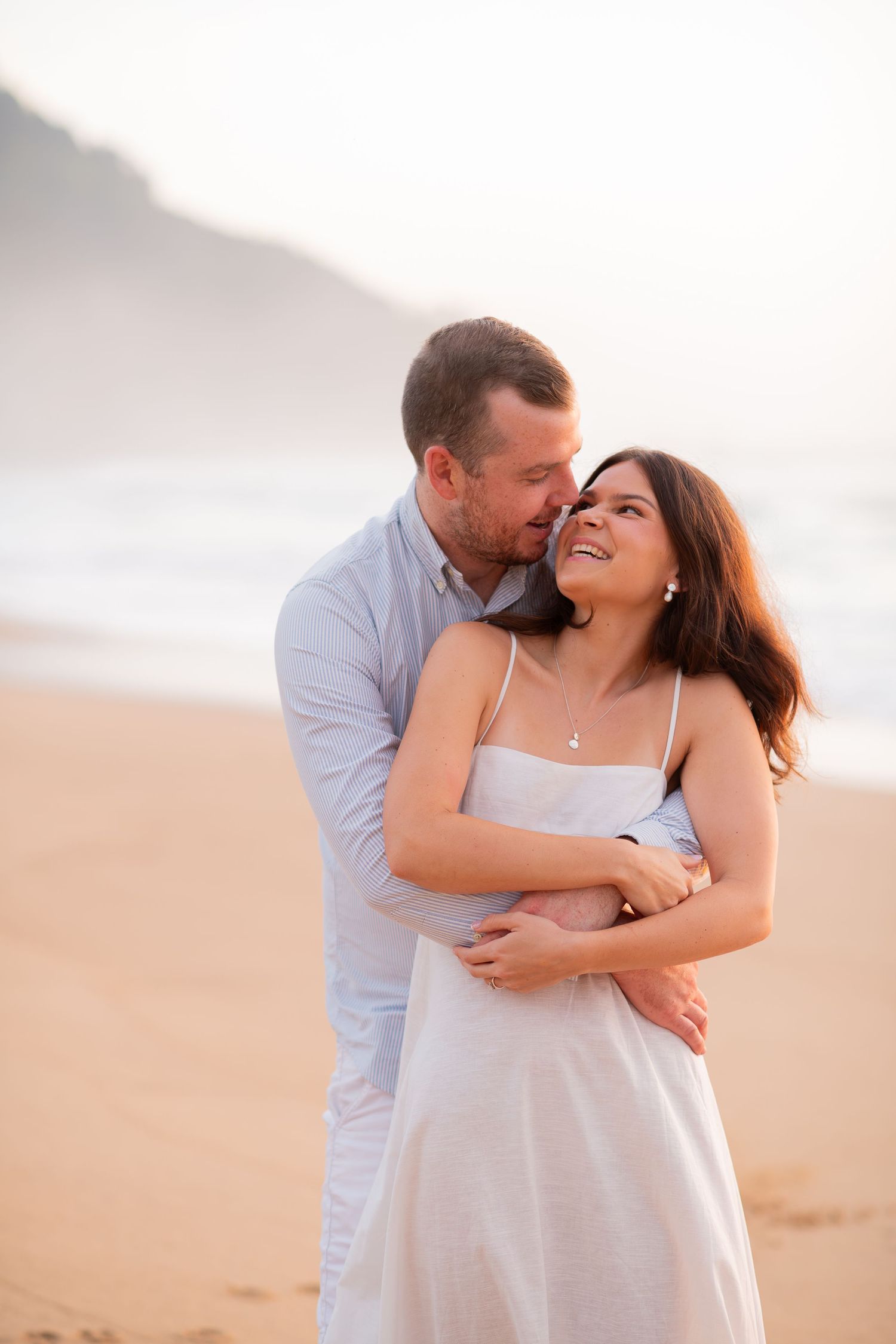 A couple embraces lovingly on a sandy beach at sunset wearing white and blue casual attire.