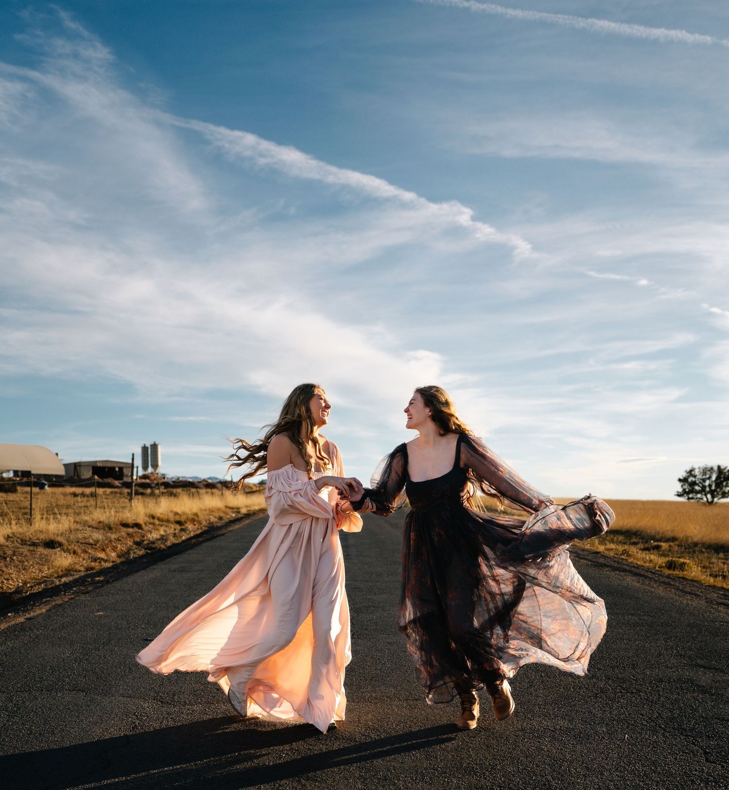 Two figures in flowing dresses dance and twirl together on an empty road at sunset with dramatic sky above.