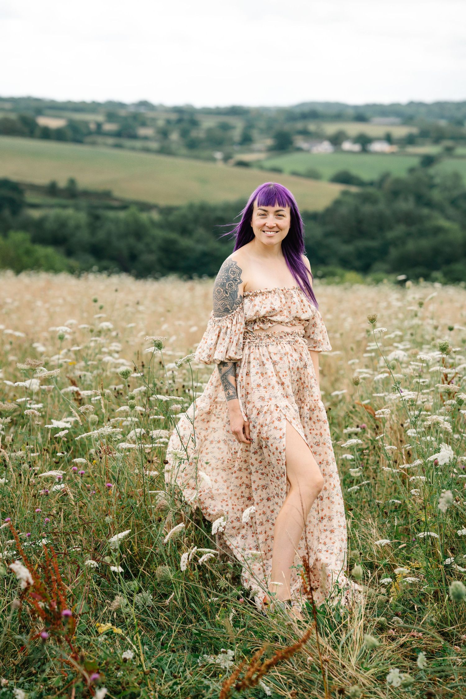A person in a floral dress stands in a field of white wildflowers and tall grass on a sunny summer day.
