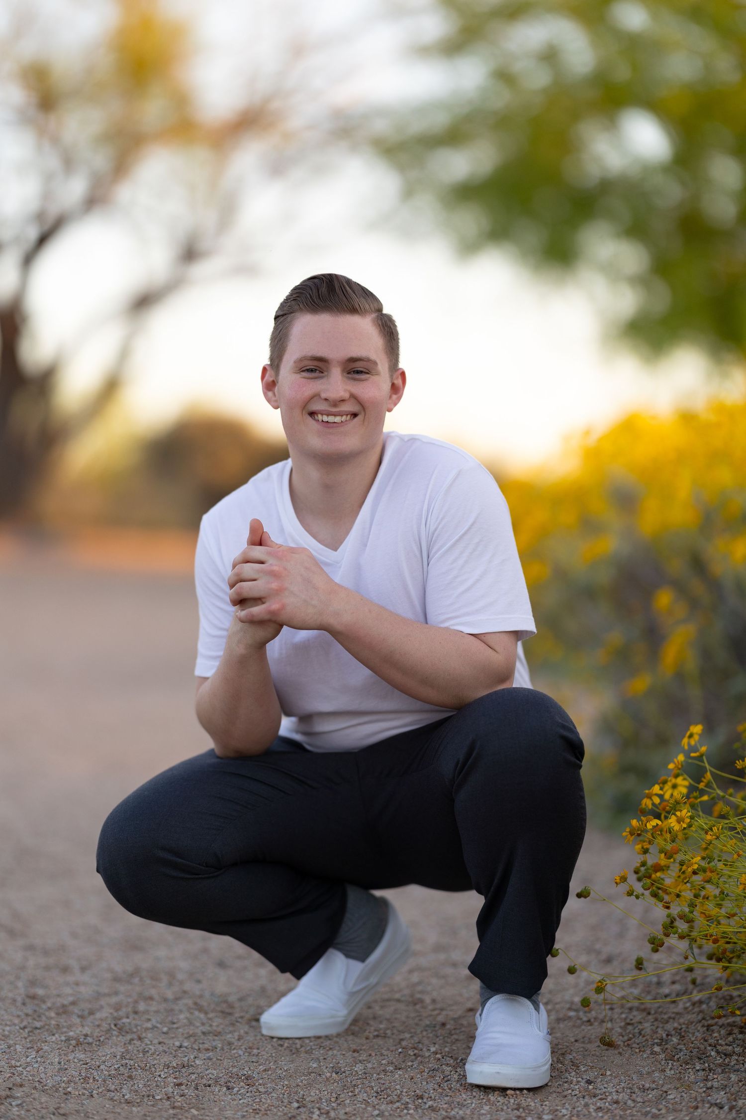 Young man in white t-shirt and dark pants crouching on a desert path, smiling, with yellow wildflowers nearby.