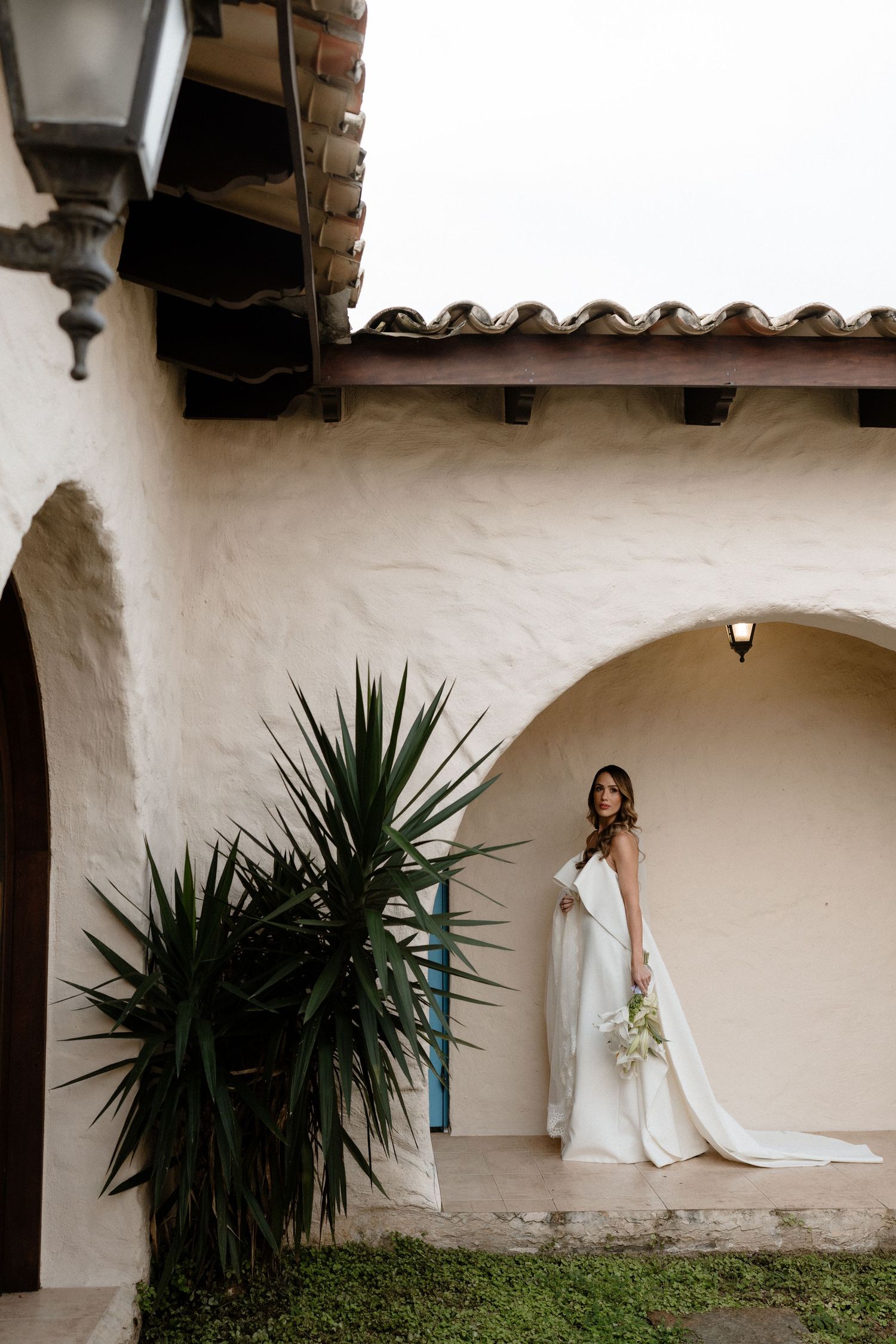 Bride in white gown holding bouquet stands in arched doorway of Spanish-style building with terracotta roof and tropical plants.