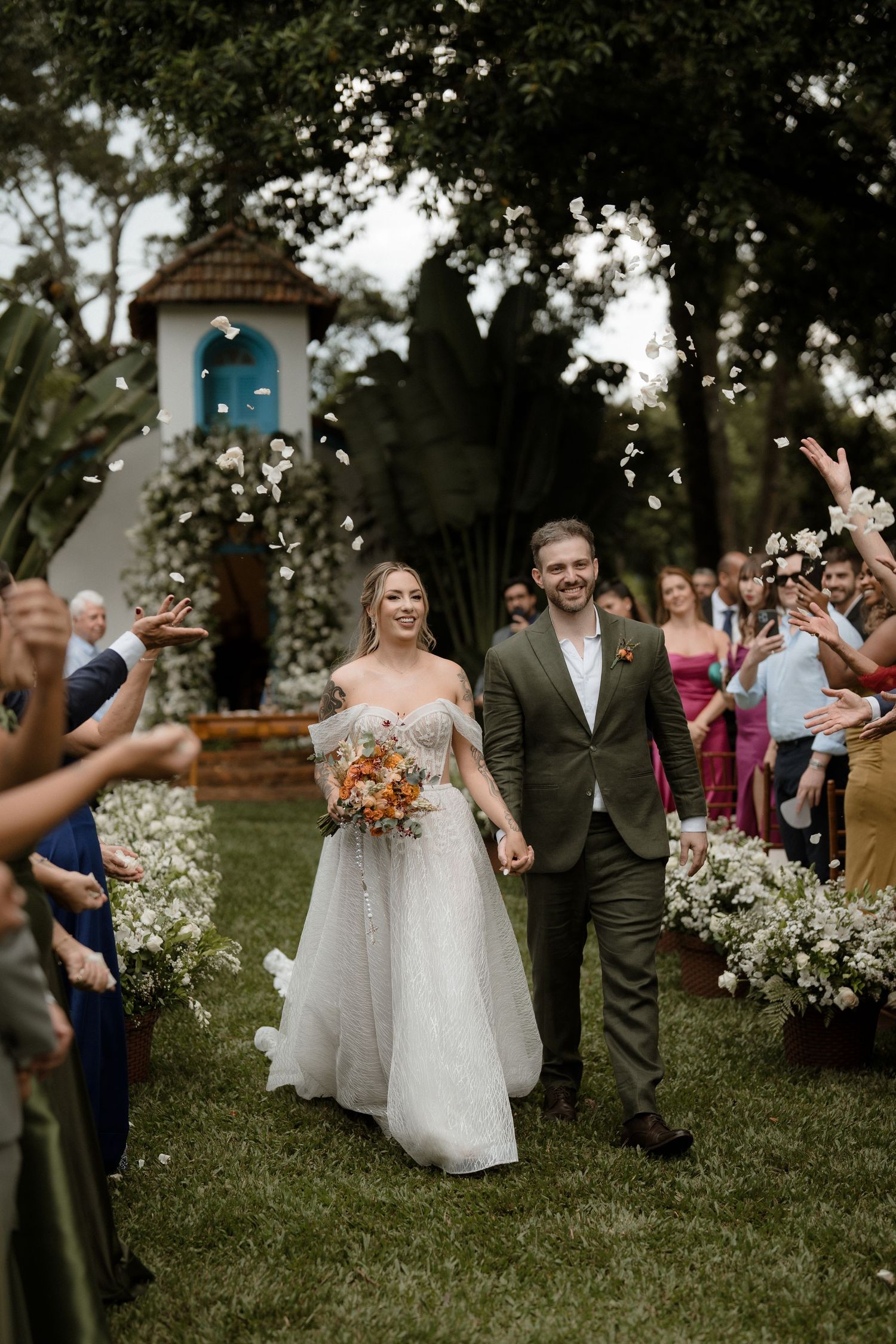 Smiling bride and groom walk down outdoor aisle as guests toss flower petals near a charming white chapel.