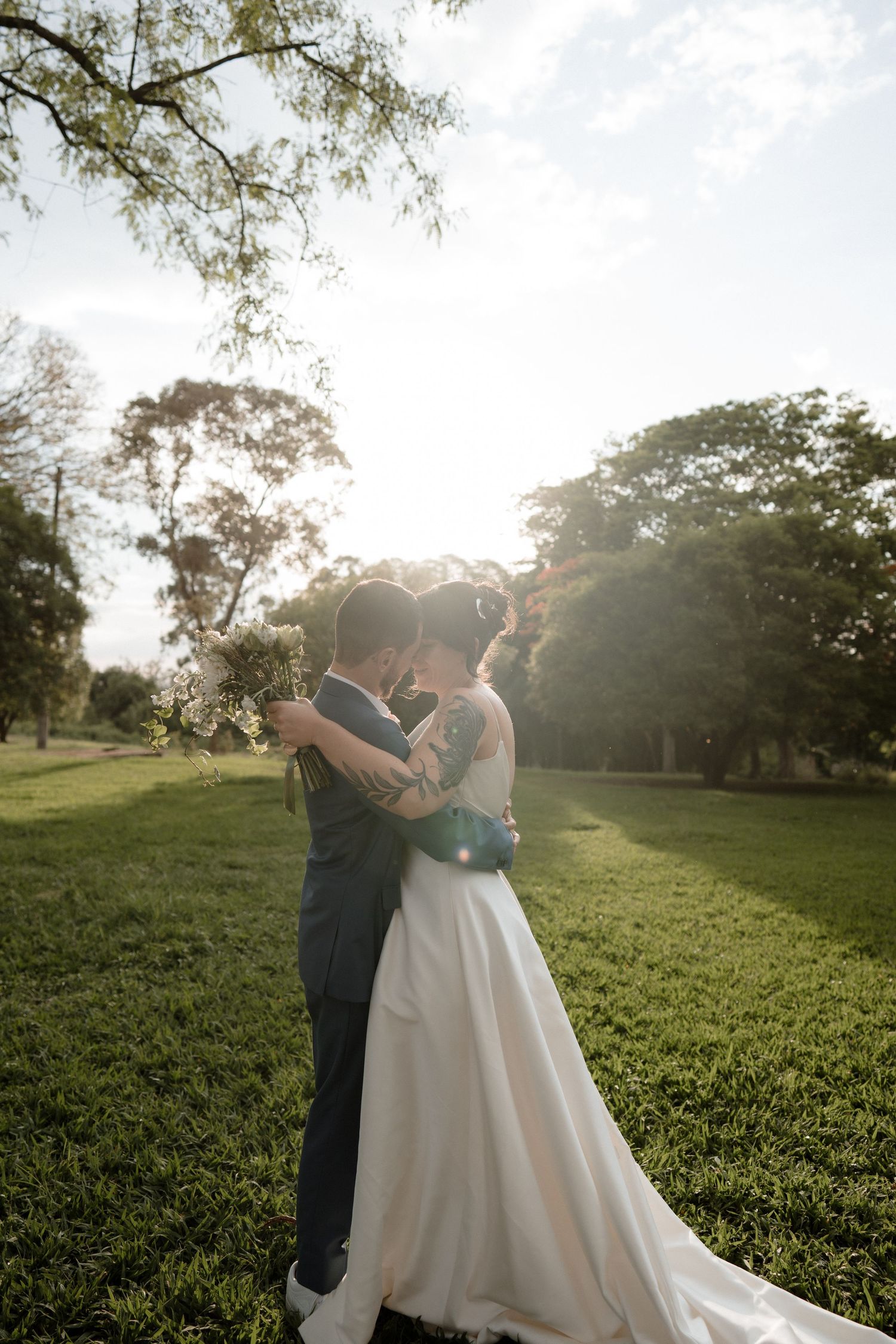 Bride and groom embracing in a sunlit park, bride in a flowing white gown, surrounded by lush green trees.