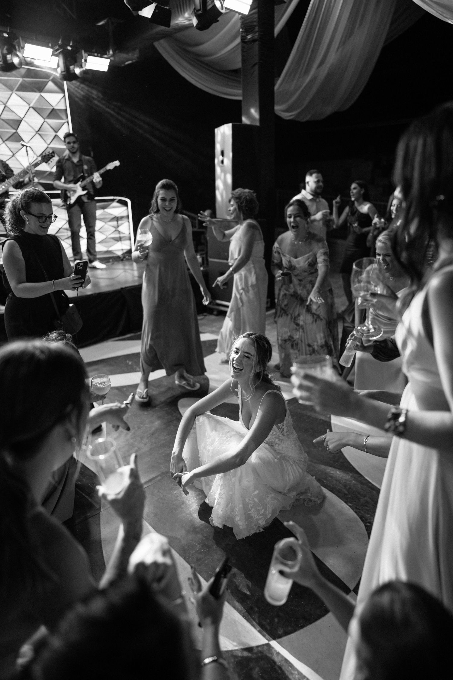 Black and white photo of a bride dancing joyfully at her wedding reception surrounded by guests and a live band.