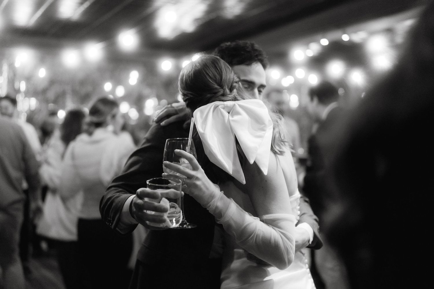 A romantic black and white moment of a couple dancing close together under glowing string lights at a celebration.