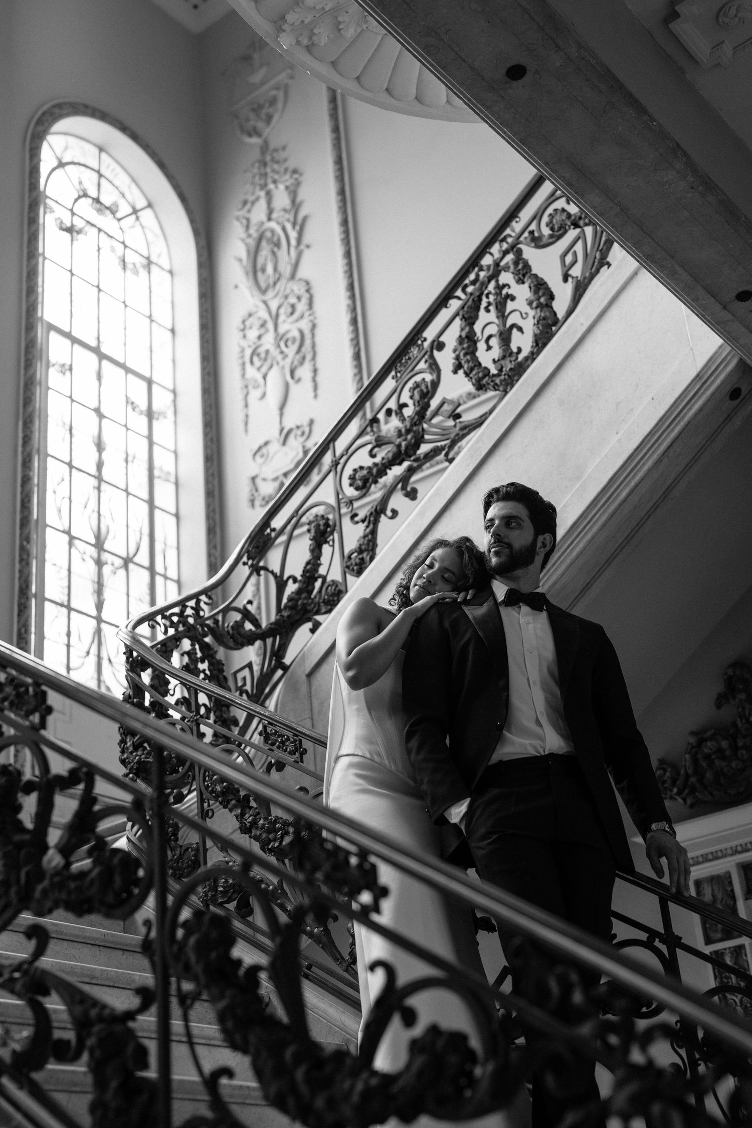 Black and white photo of elegant bride and groom descending ornate staircase in grand venue with decorative ironwork.