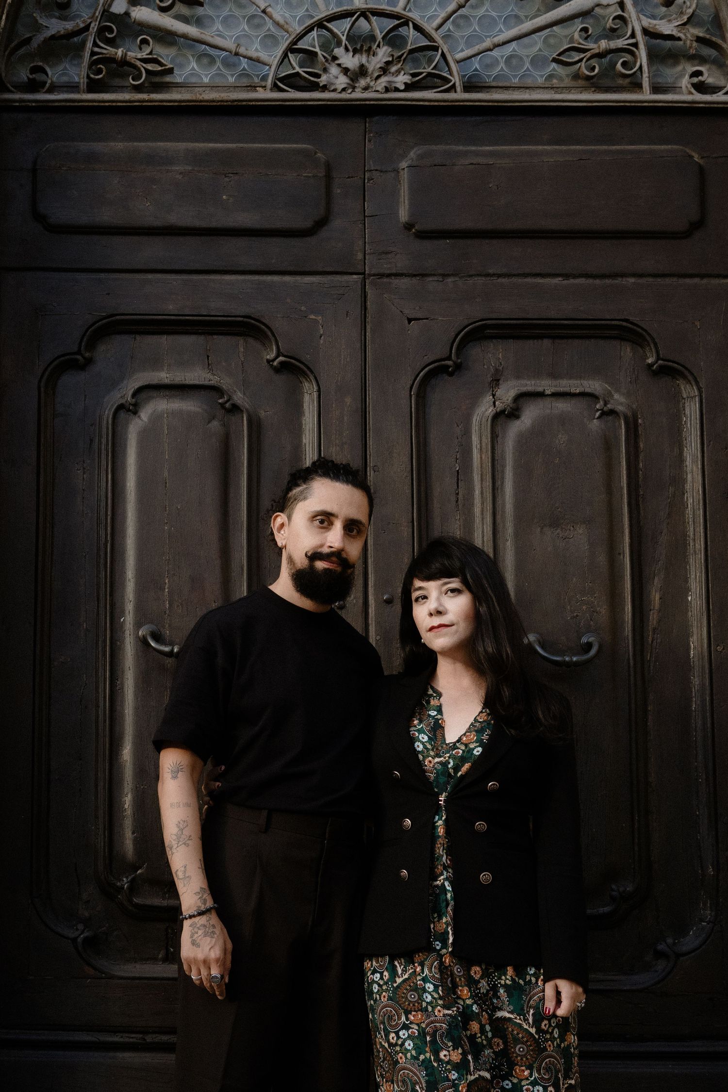 A bearded man with tattoos and a woman in a floral dress pose before ornate dark wooden doors.