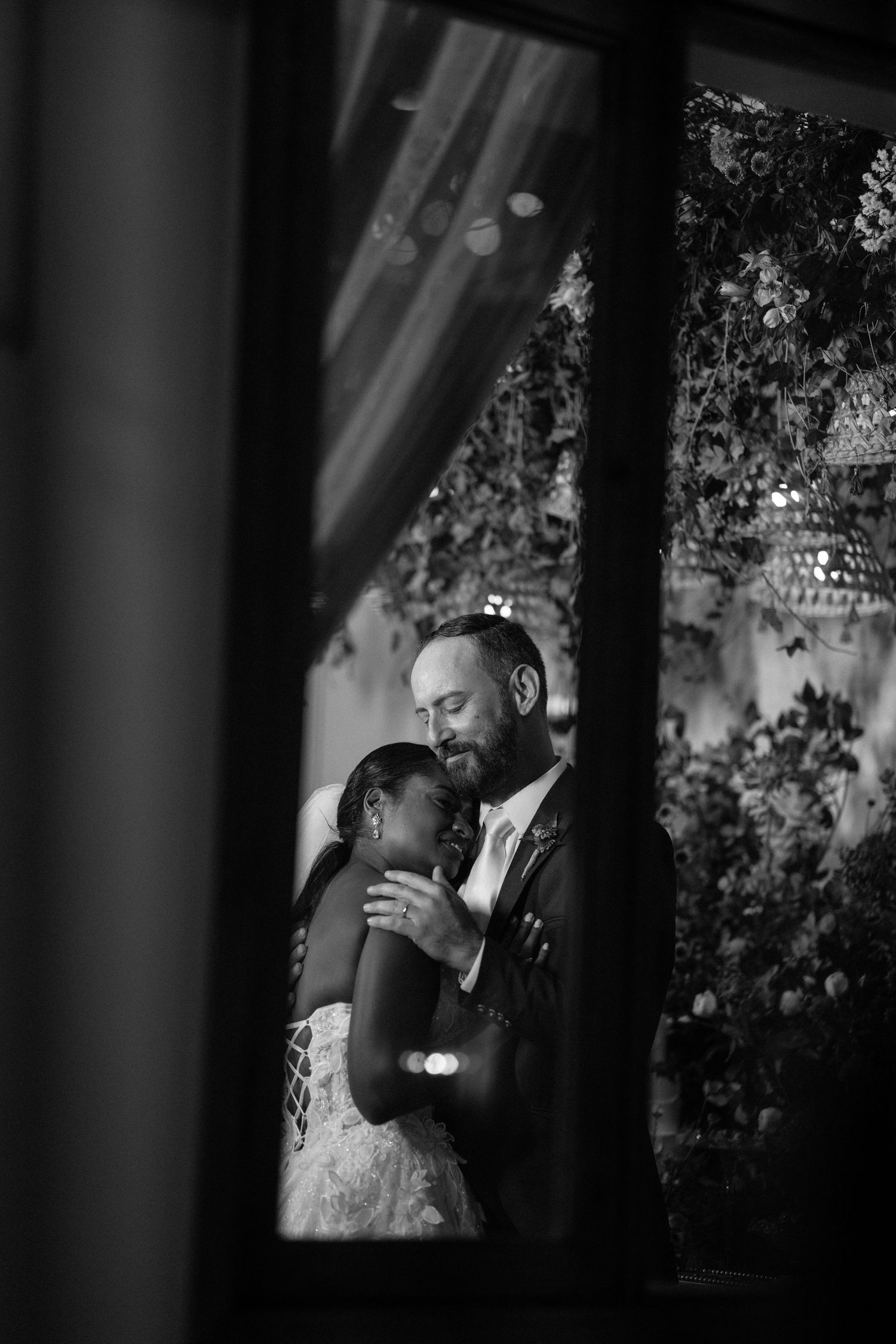 Black and white wedding photo of bride and groom embracing tenderly, framed by a window with floral backdrop.
