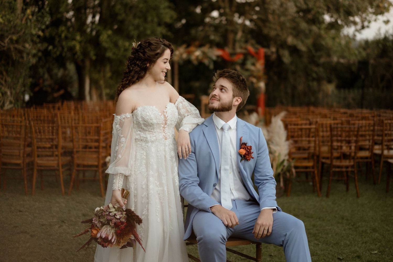 A romantic outdoor wedding scene with a couple in white dress and blue suit sitting on wooden chairs in a garden setting.