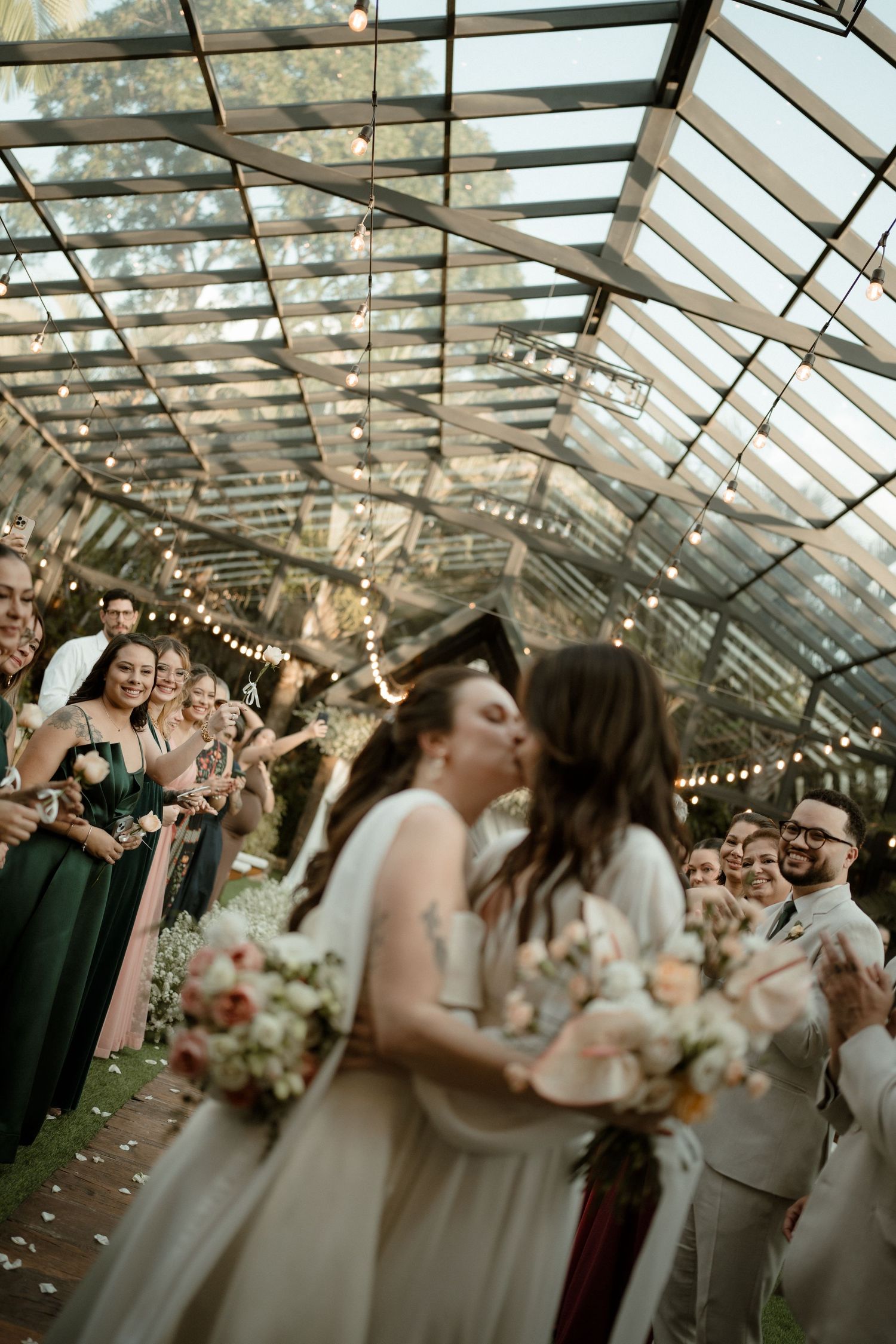 Wedding celebration inside a greenhouse venue with string lights and guests gathered for a joyful moment.