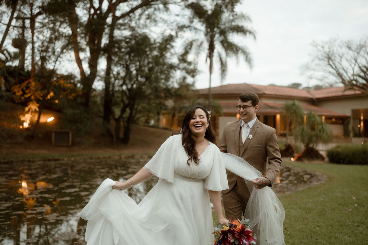 Couple in wedding attire poses together outdoors during golden hour at a scenic venue with palm trees.