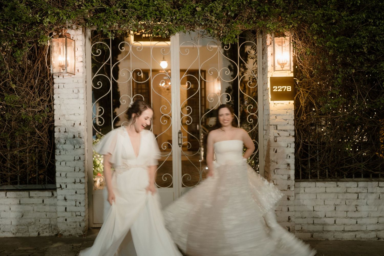 Two people in elegant white gowns pose together in front of a white brick wall with glowing outdoor sconces.