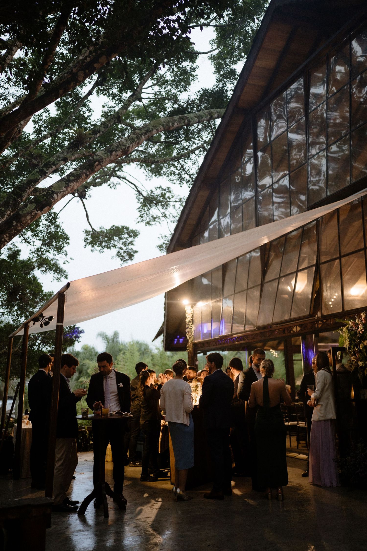 Elegant outdoor evening wedding reception with guests mingling under large trees and warm string lights near a modern venue.