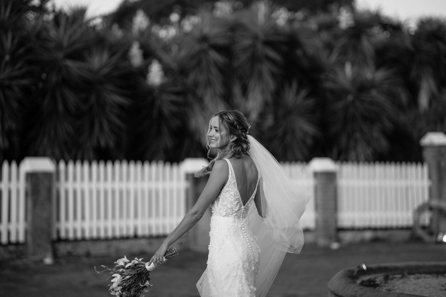 Black and white photo of a bride in a backless wedding dress holding flowers against a white picket fence and palm trees.
