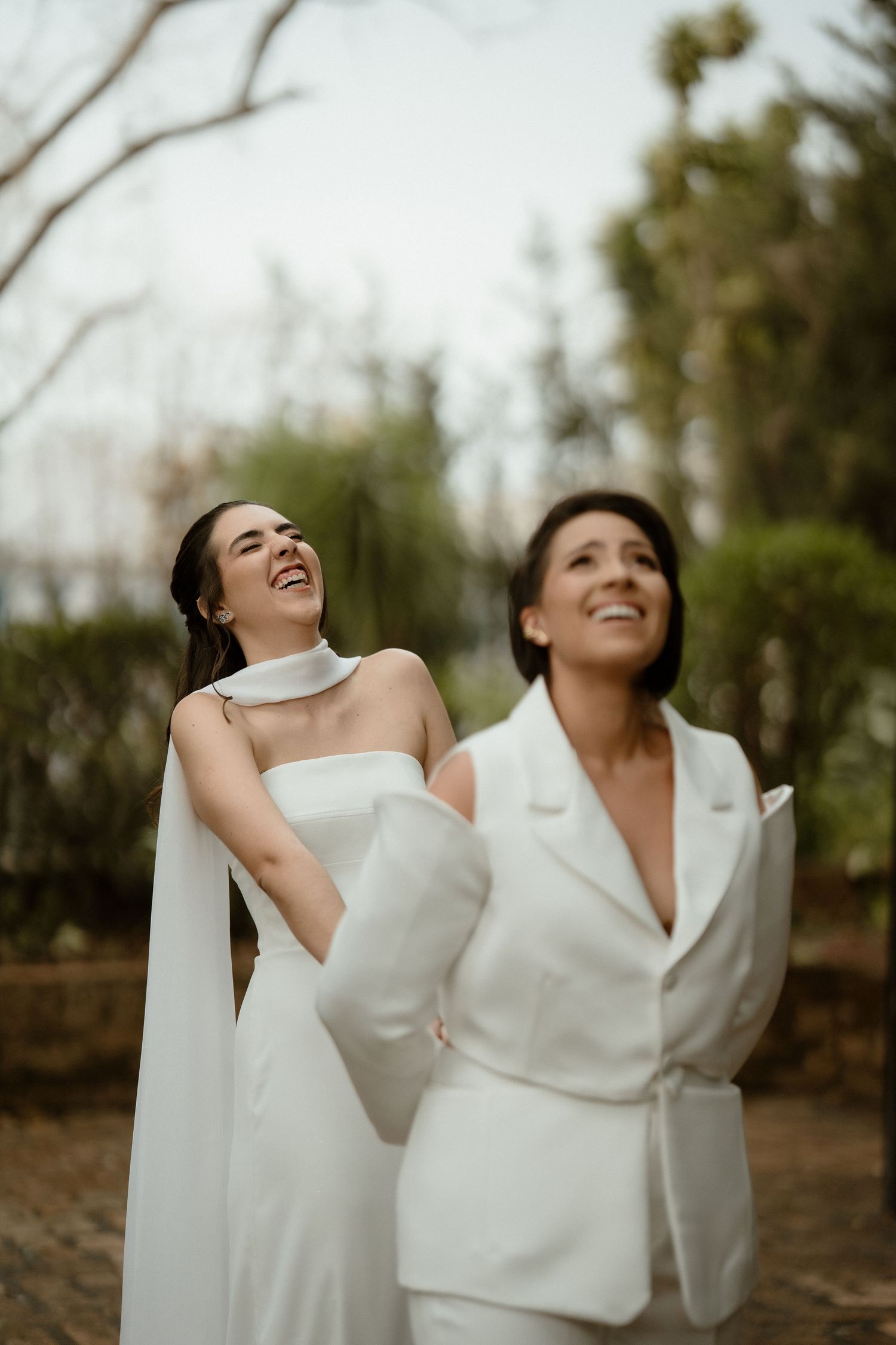 Two people in elegant white wedding attire share a joyful candid moment outdoors in a natural setting.