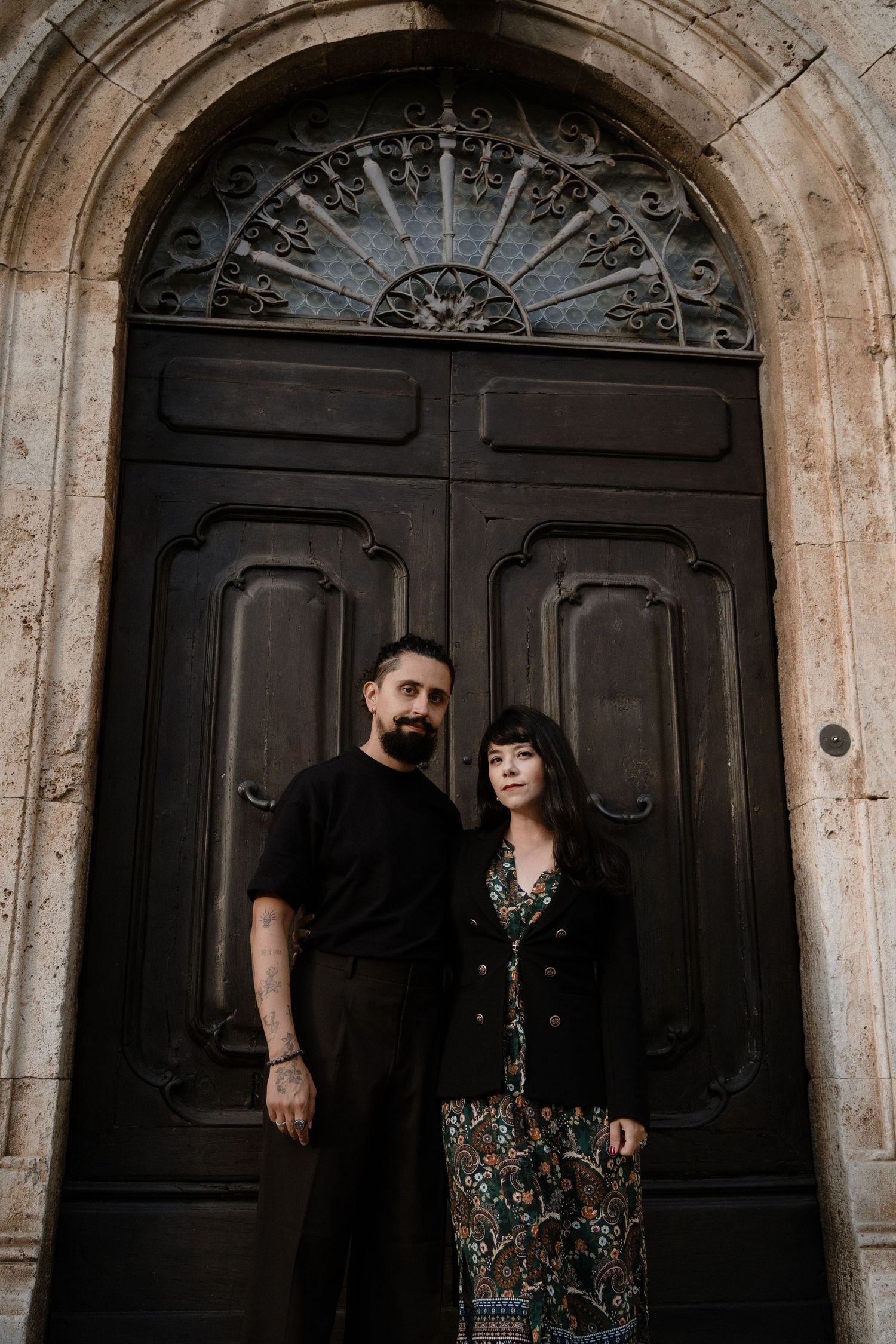 Couple standing before ornate dark wooden arched door with decorative ironwork in historic stone building.