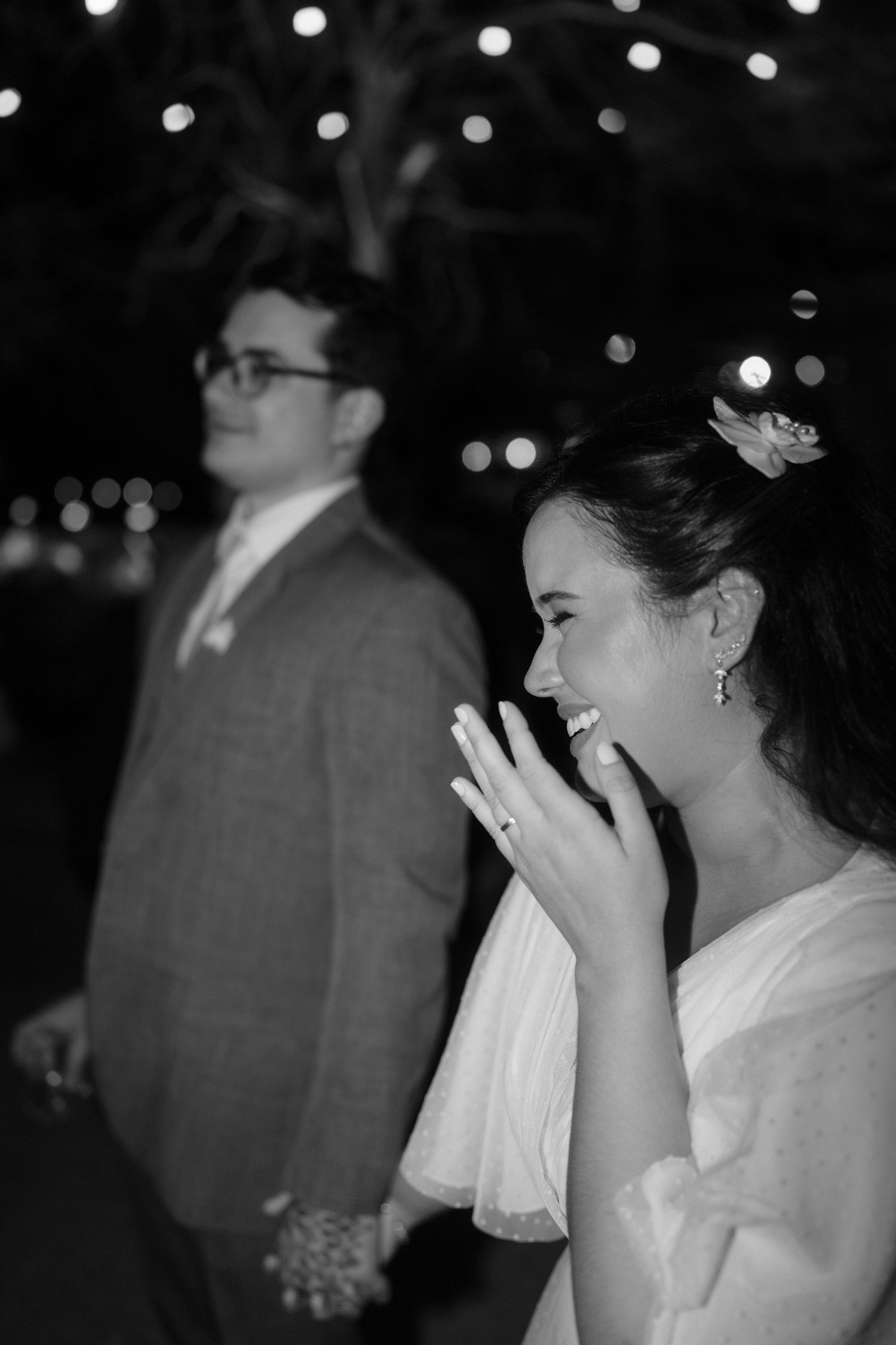 Black and white wedding photo of smiling bride showing ring while groom stands behind her, bokeh lights in background.