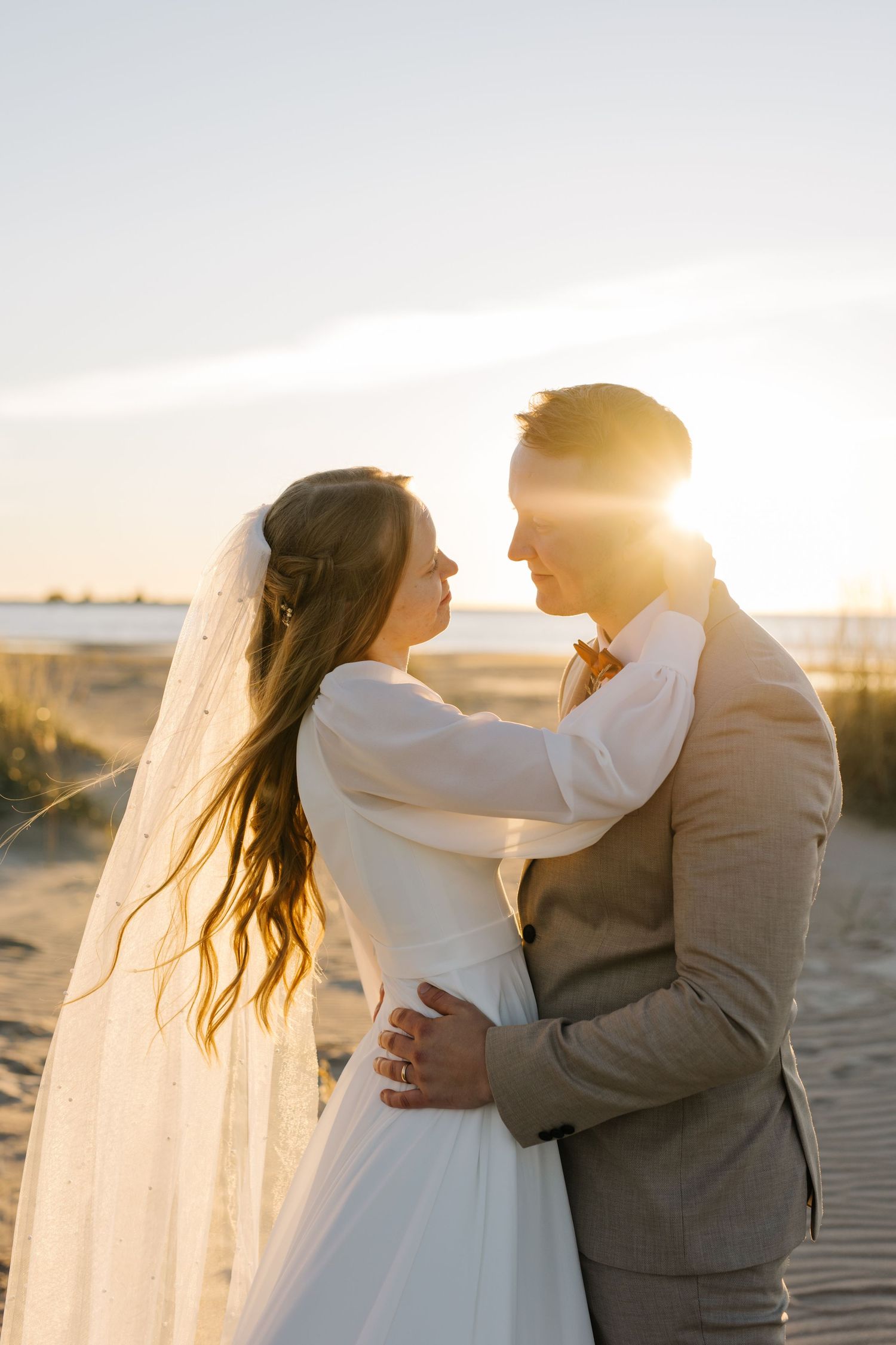 A couple in wedding attire embraces on a beach at sunset, their silhouettes backlit by golden sunlight streaming through clouds.