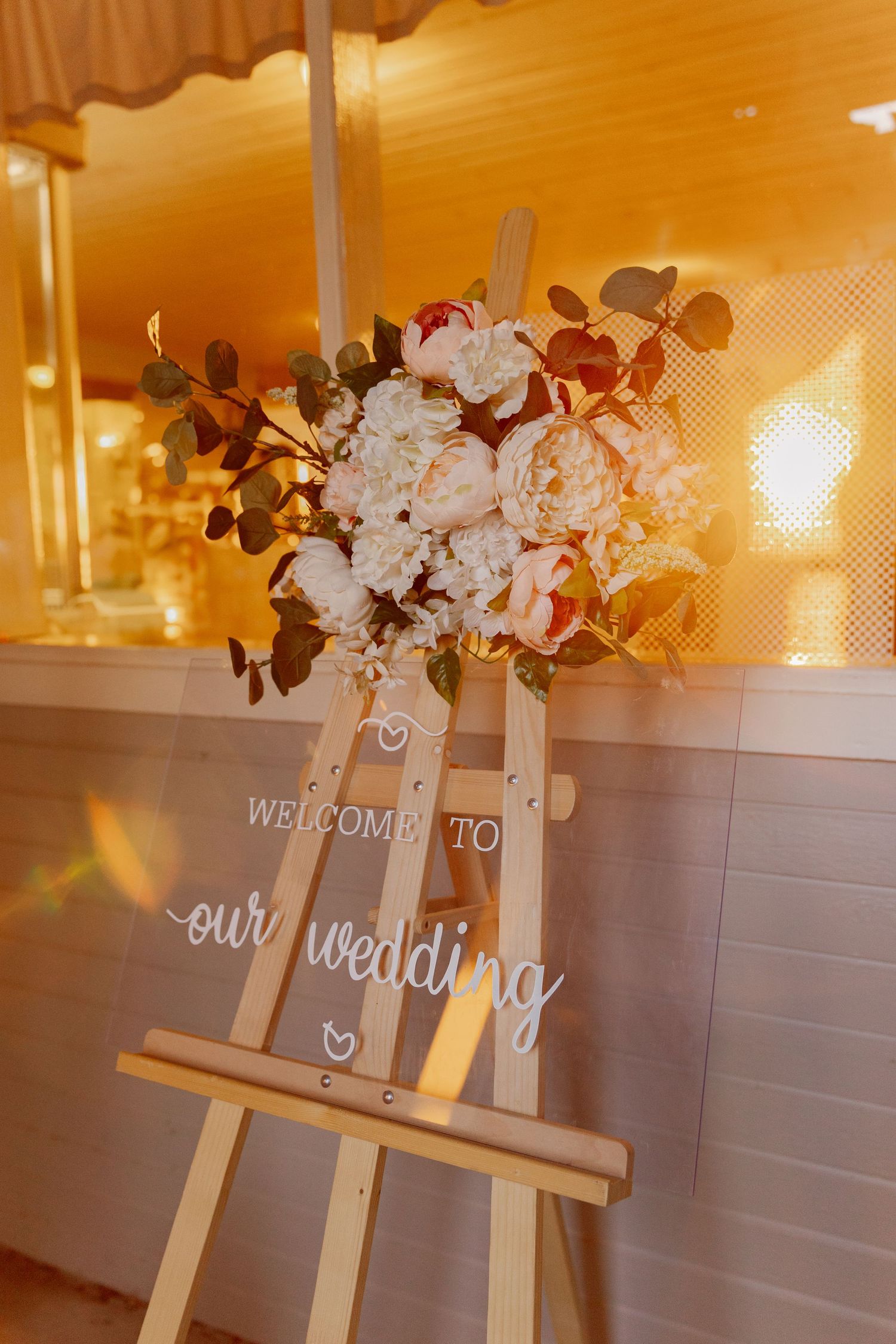 A wedding welcome sign on an easel decorated with white and pink flowers in warm lighting.