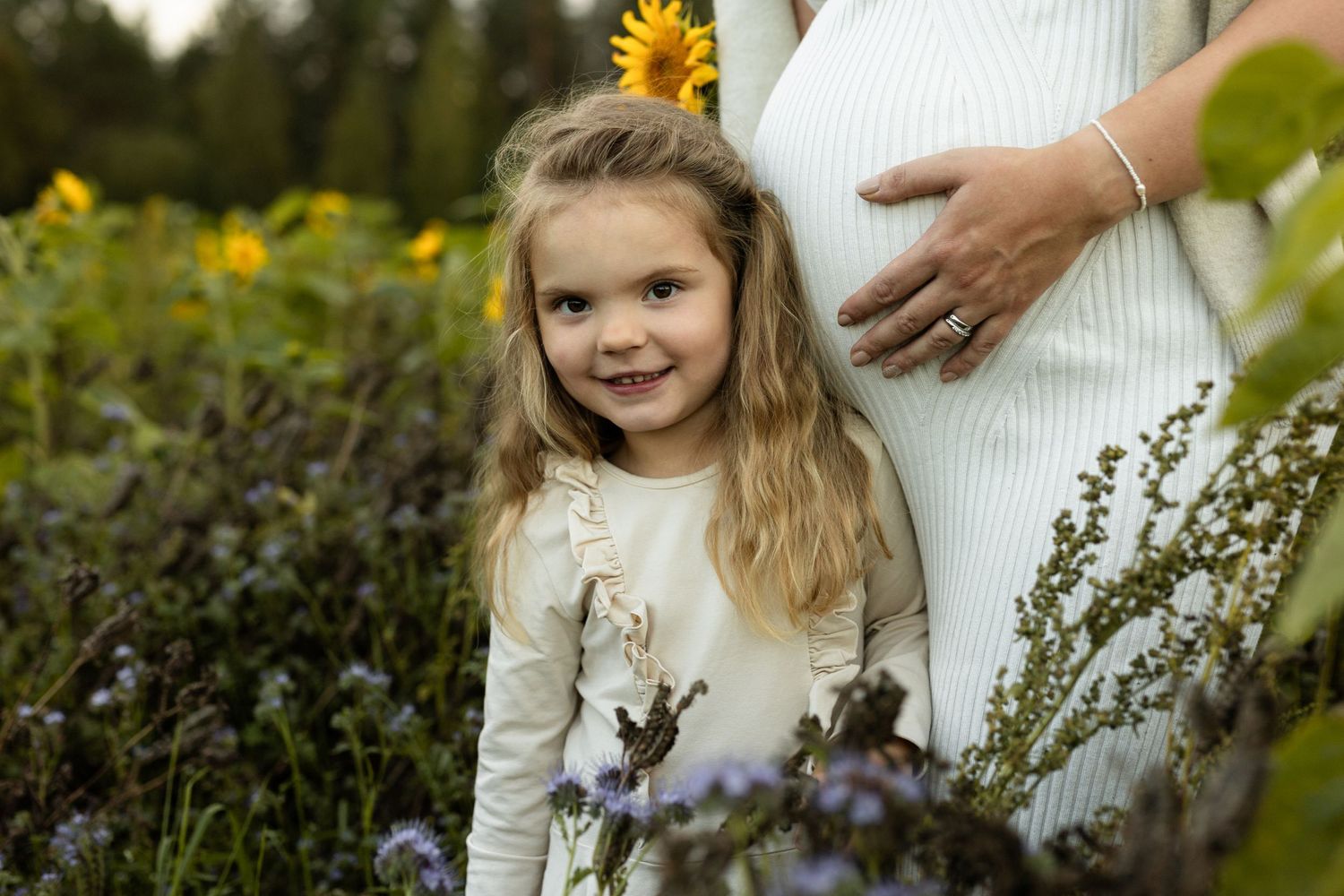 Child standing beside pregnant belly in field of yellow flowers and purple wildflowers.