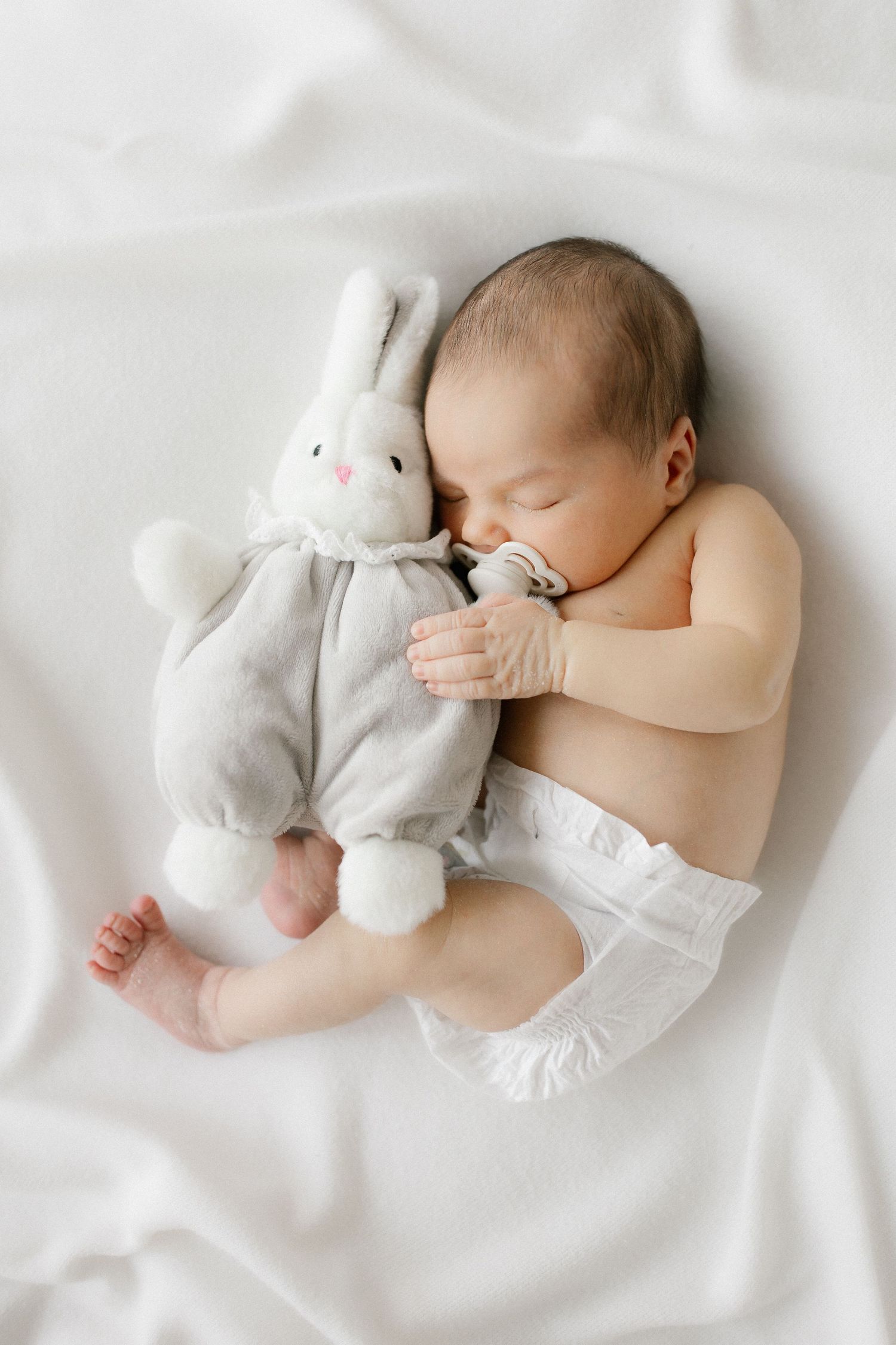 Newborn baby sleeping peacefully in a diaper, cuddling a soft gray bunny stuffed animal on a white blanket.