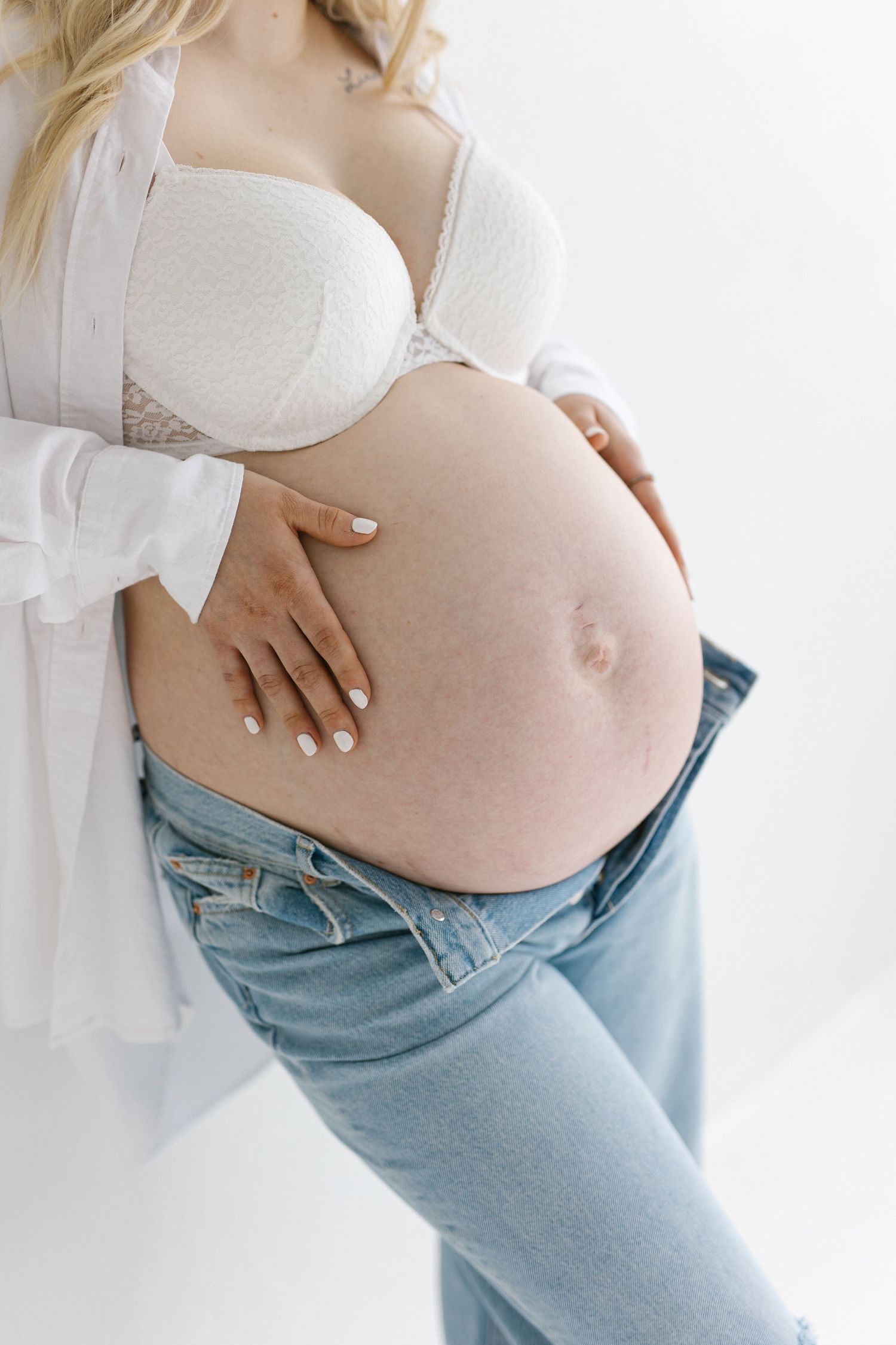 Pregnant woman in white bra and jeans showing her bare belly in a series of intimate maternity photos.